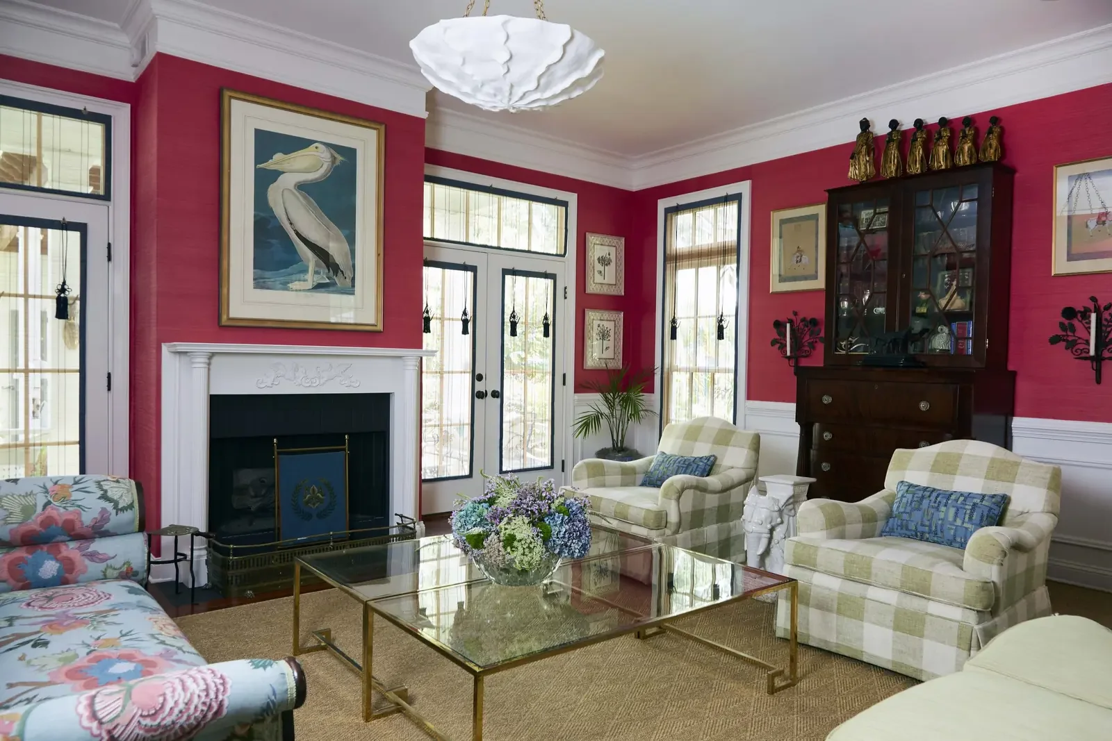 Living room with red walls, white trim, and large windows. Features checked armchairs, a glass coffee table with a flower arrangement, a dark wood display cabinet, and framed artwork including a large framed pelican.
