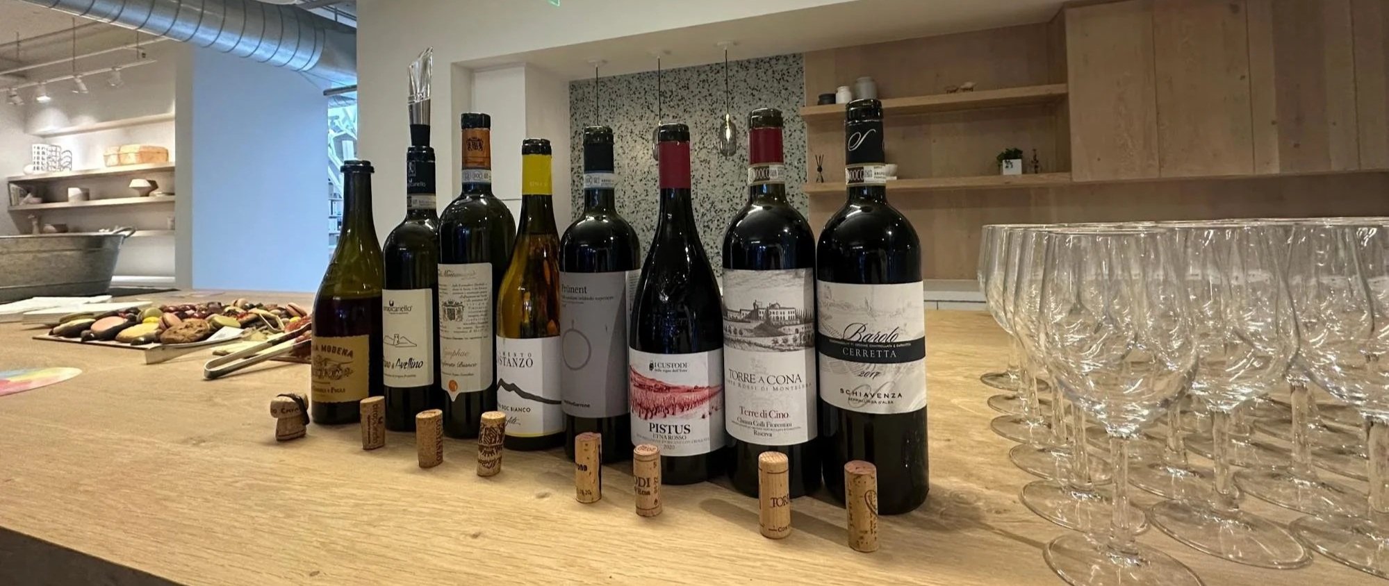 Lineup of seven wine bottles with corks in front, next to empty wine glasses on a wooden counter in a modern, minimally decorated kitchen or wine bar.