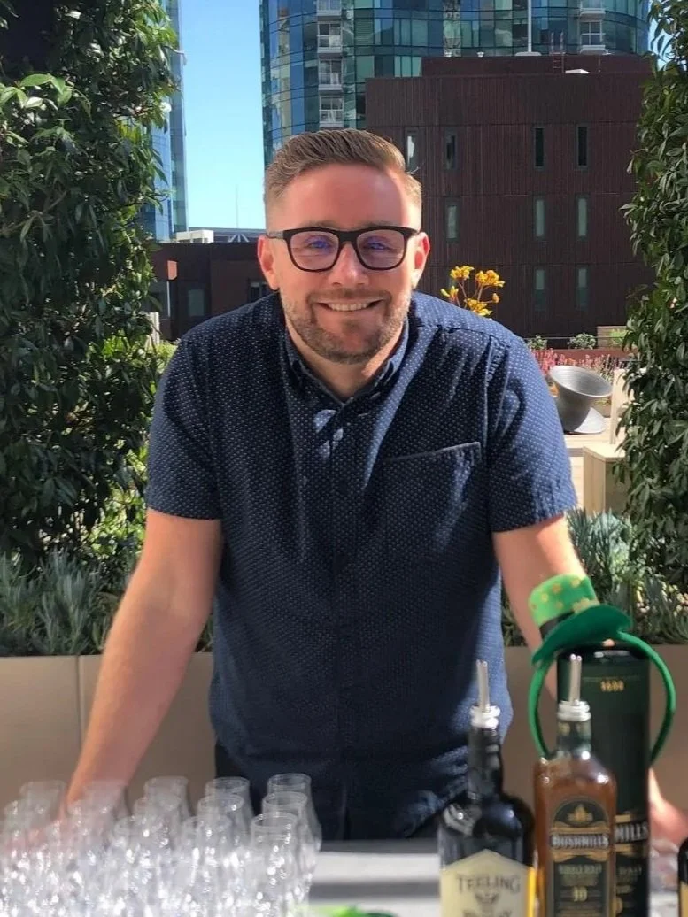A man with glasses and a beard smiling, standing behind a table with bottled cocktail ingredients and empty glasses, outdoors with modern buildings and greenery in the background.