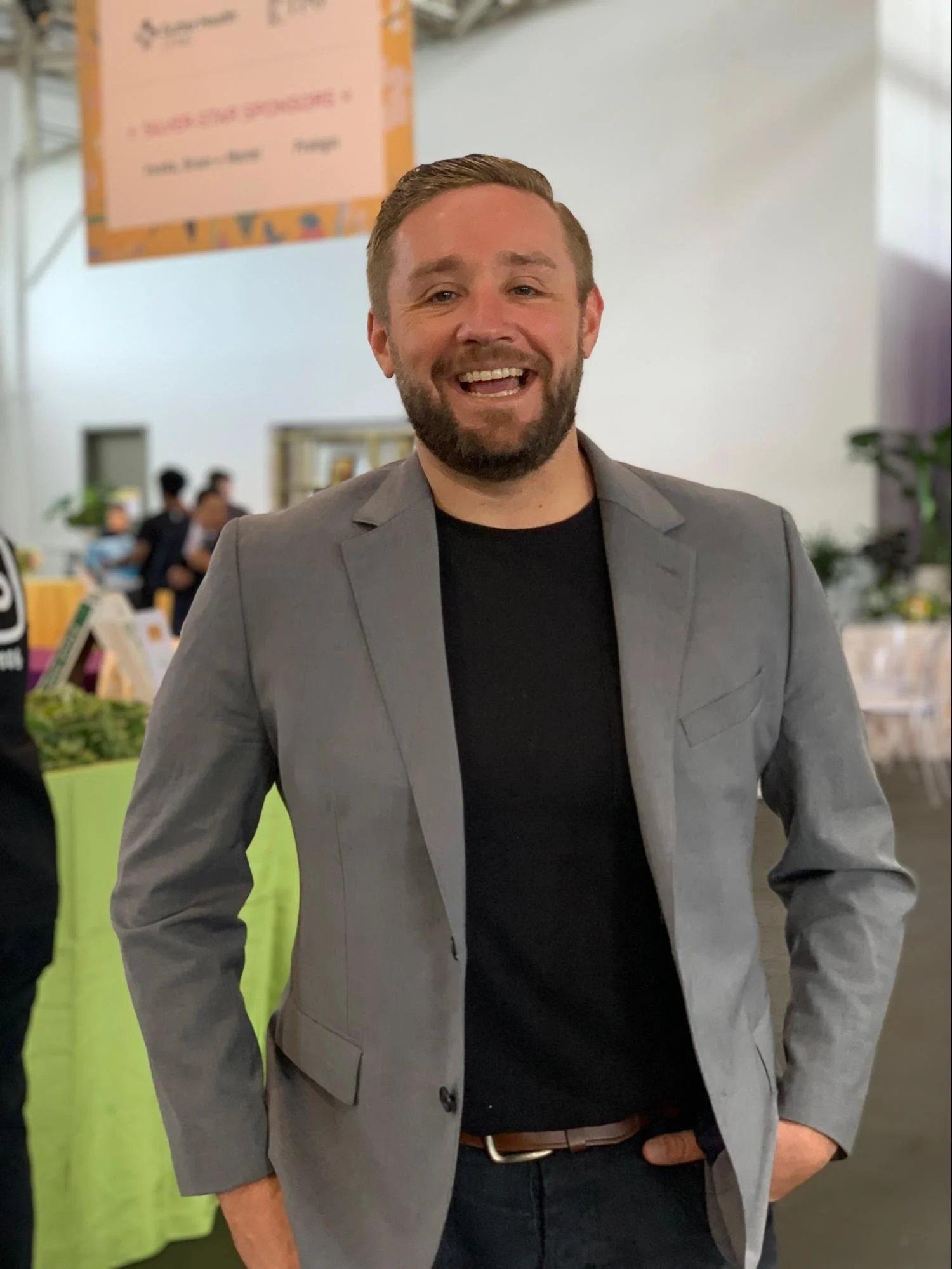 A smiling man in a gray blazer and black shirt standing indoors at an event. Background includes other people and a table with produce.