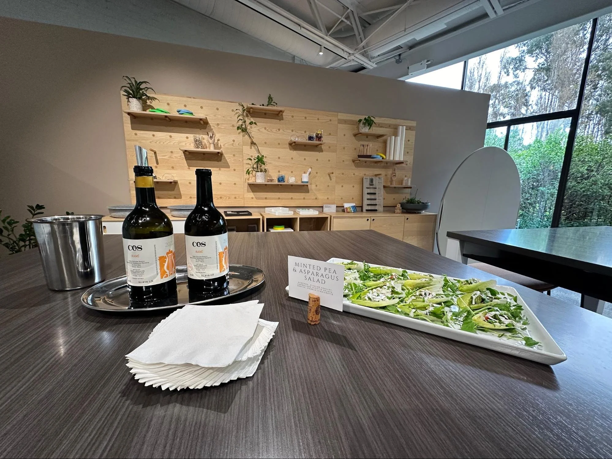 A table with two bottles of wine, a silver tray, a stack of white napkins, and a large rectangular dish of pea and asparagus salad with a label. In the background, a wooden wall with shelves holds plants, books, and decorative items, and large windows reveal a wooded outdoor scene.