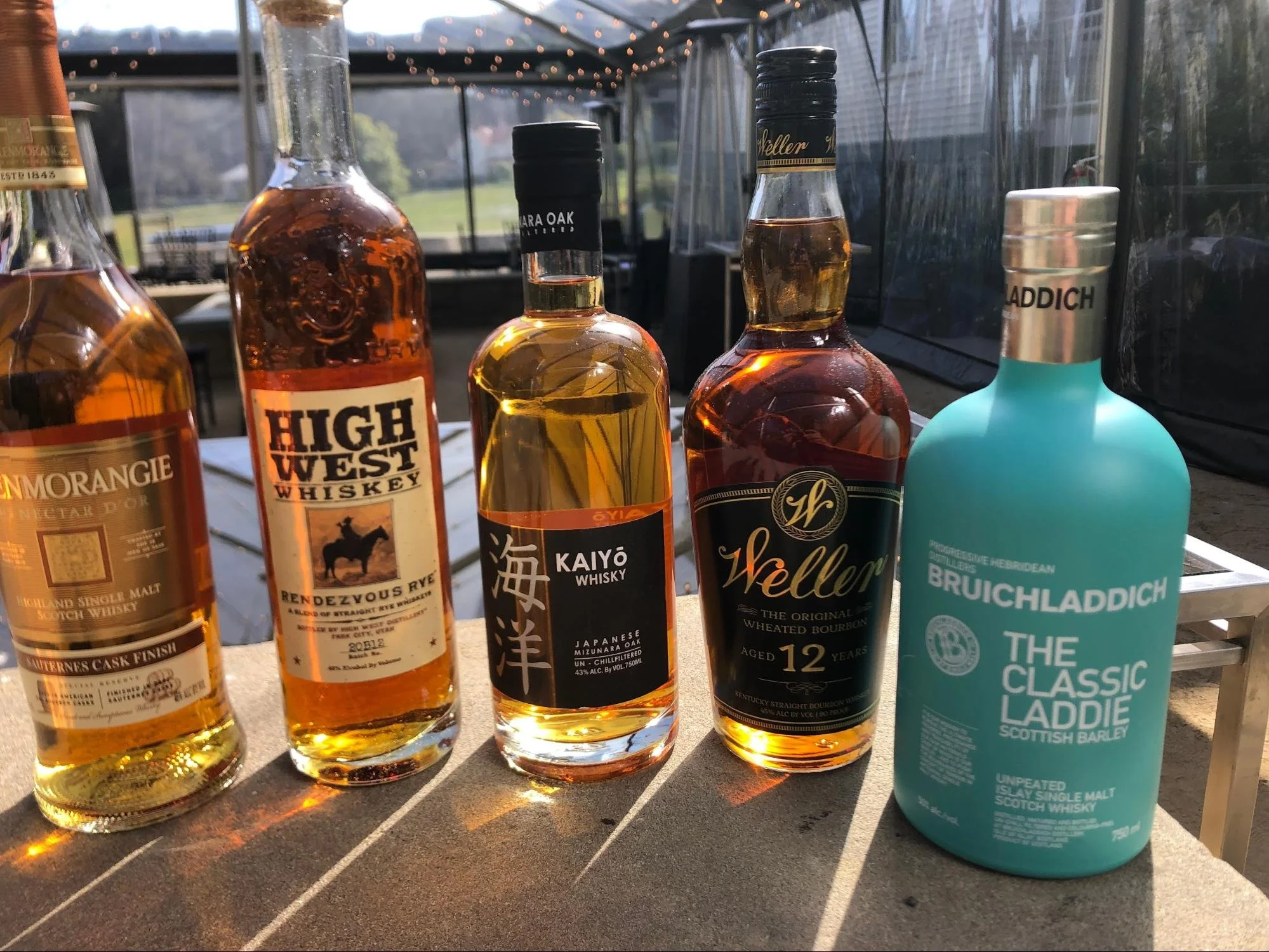 Five bottles of whiskey and scotch whisky sitting on a table outdoors, with a blurred background of string lights and a windowed structure.