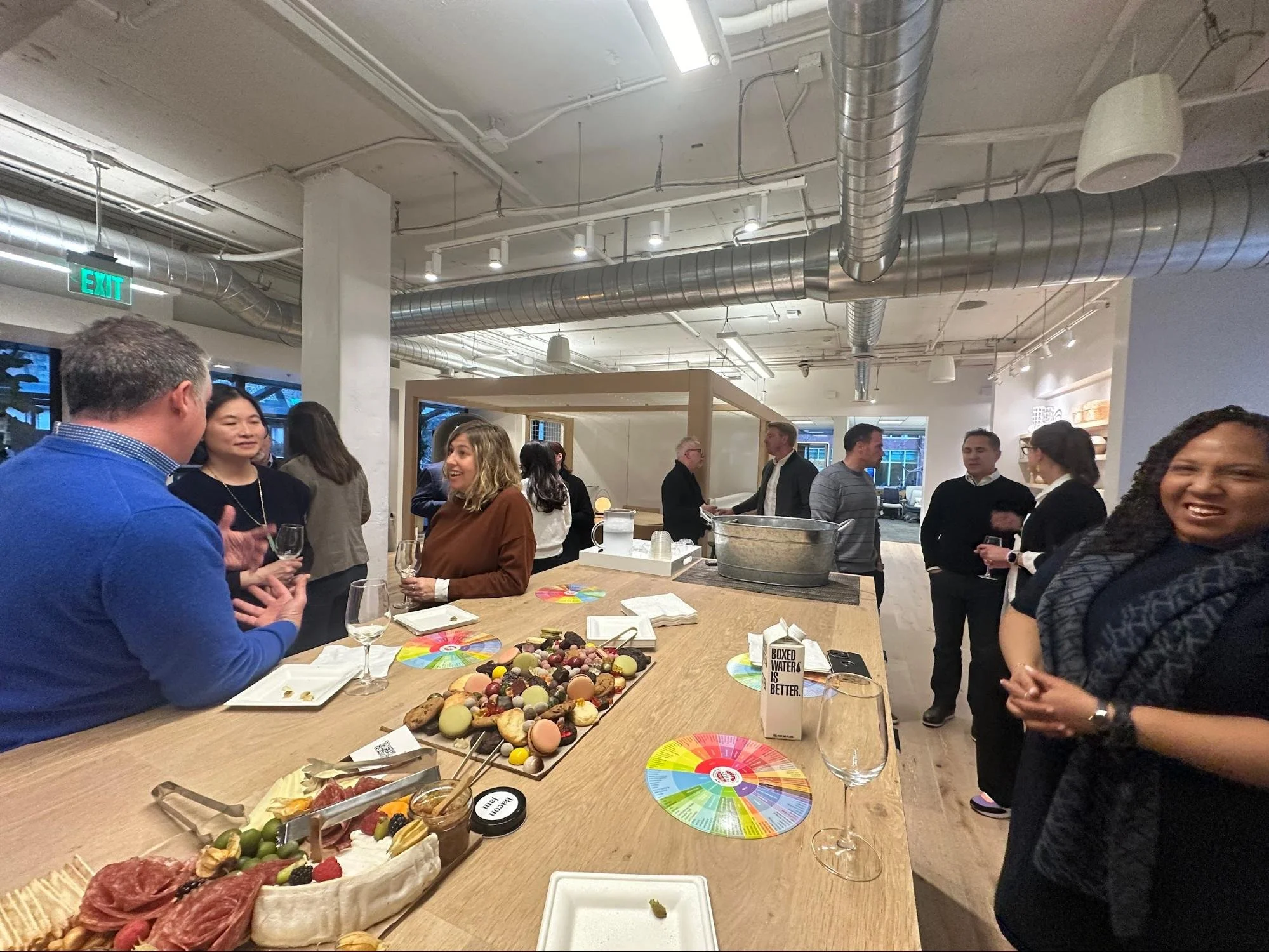 People socializing at a gathering with a food and drink table, including a cheese platter, chocolates, and wine glasses, in a modern indoor space.