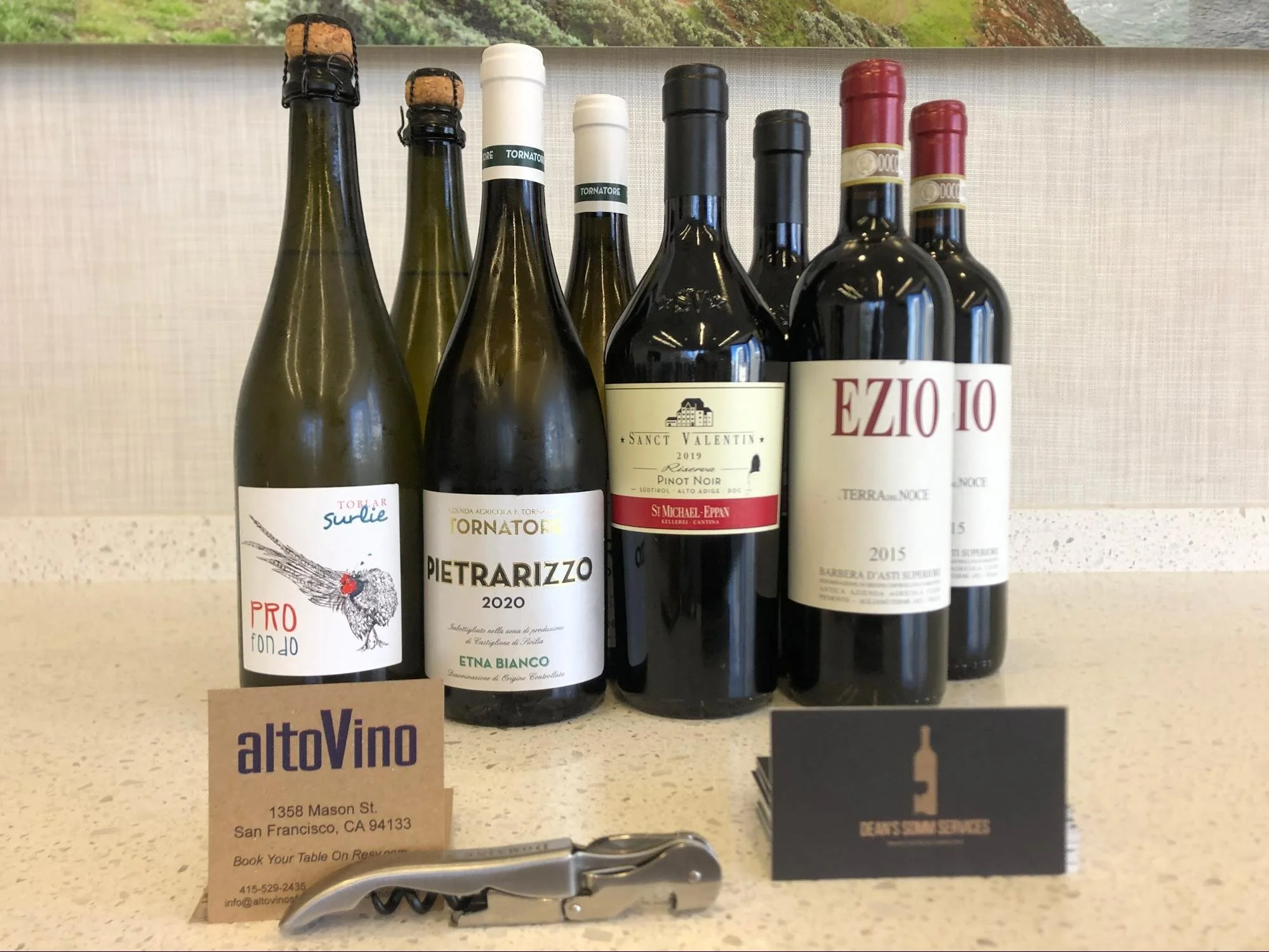 Six bottles of wine with labels facing forward on a white countertop, with business cards and a corkscrew in front. The background appears to be an outdoor or indoor setting with a window showing trees. The bottles include sparkling wine with corks, and various red and white wine bottles.