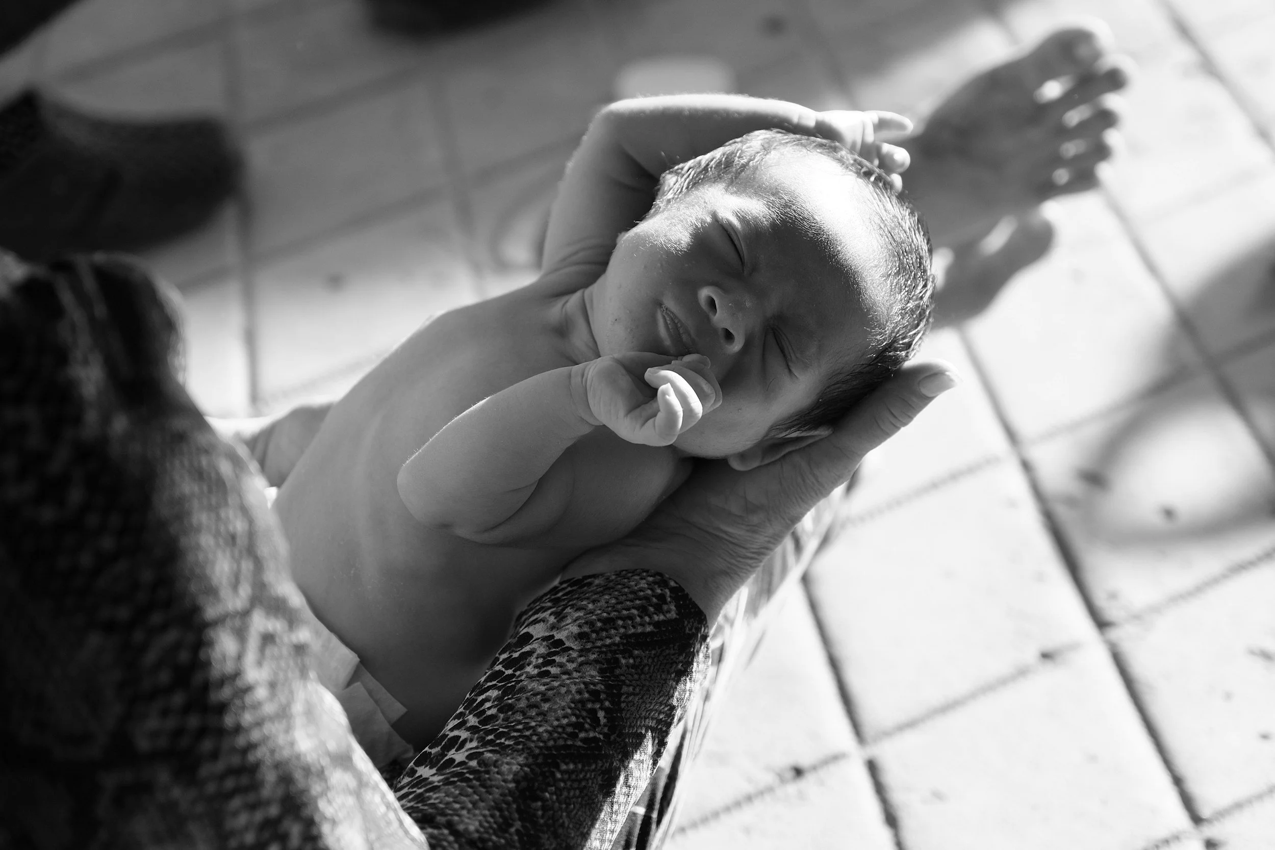 Black and white image of a peaceful newborn baby cradled in adult hands. The baby's eyes are closed, and its expression is serene. Sunlit brick floor in the background.