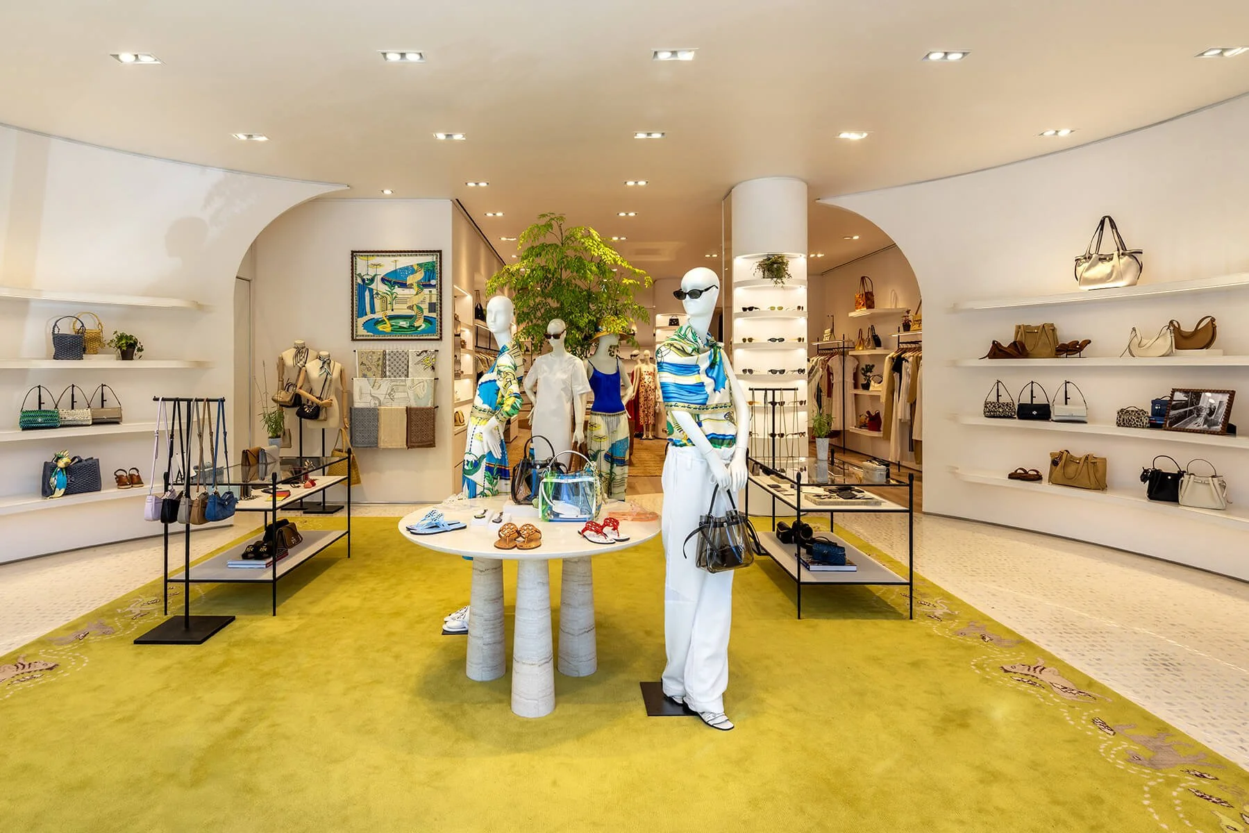 Interior of the Tory Burch Americana Manhasset store, with printed summer looks on mannequins, a chartreuse rug, and handbags along curved wall shelving

