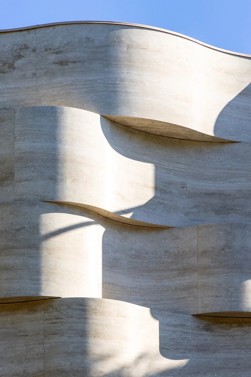 Close detail of the sculpted travertine facade at the Tory Burch Americana Manhasset store, with soft tree shadows across the curved stone
