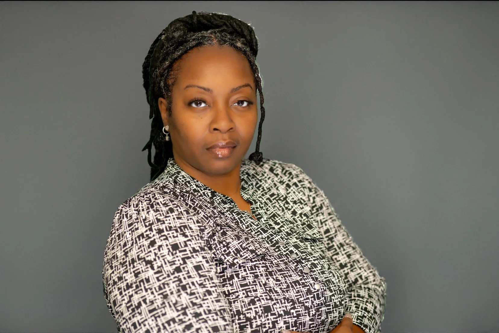 Portrait of an African American woman with dreadlocks, wearing a black and white patterned blouse, standing against a gray background.