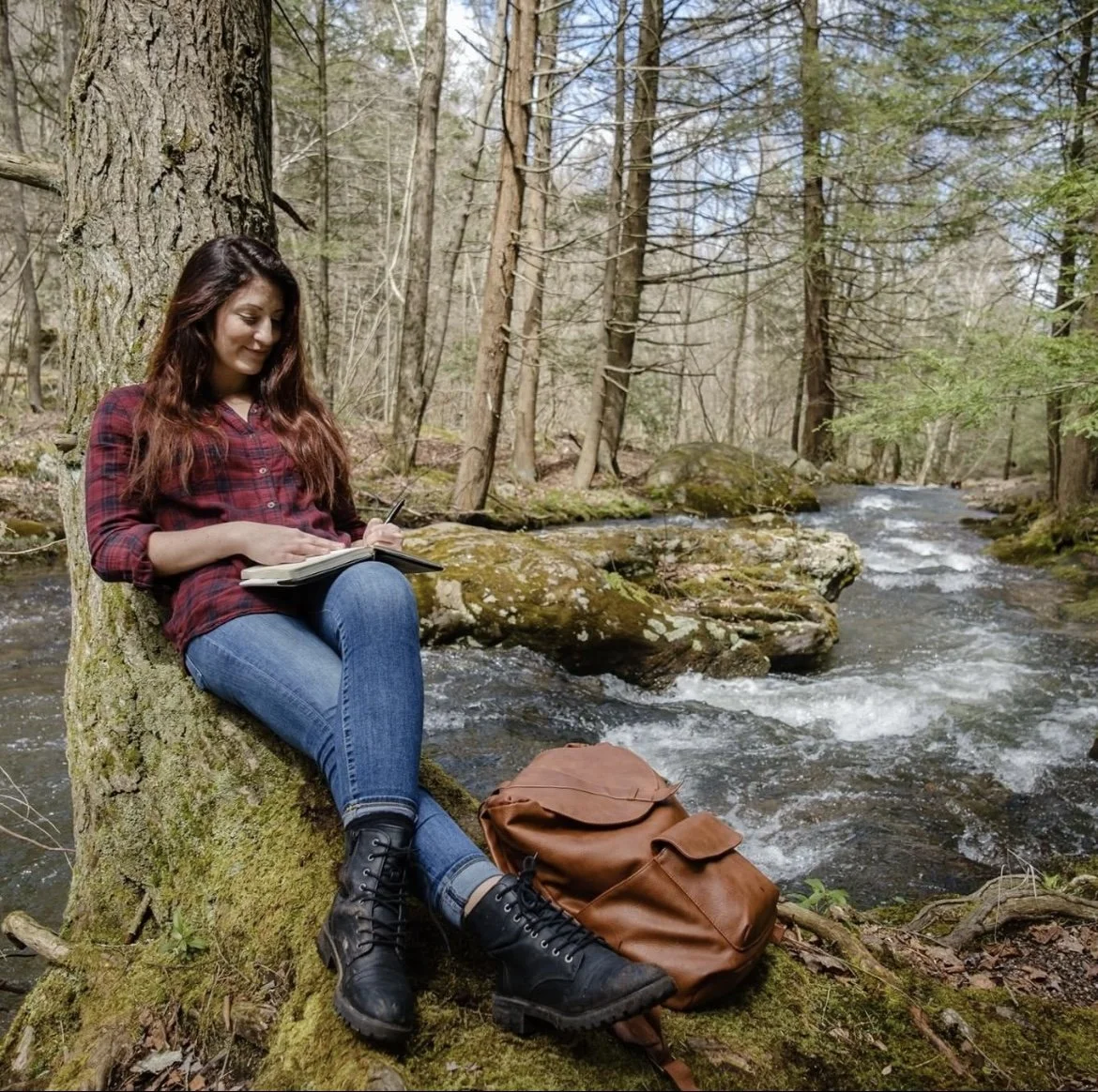 A young woman in a red plaid shirt and jeans sitting on a moss-covered tree trunk next to a stream in a forest, writing or drawing in a notebook, with a brown backpack on the ground nearby.