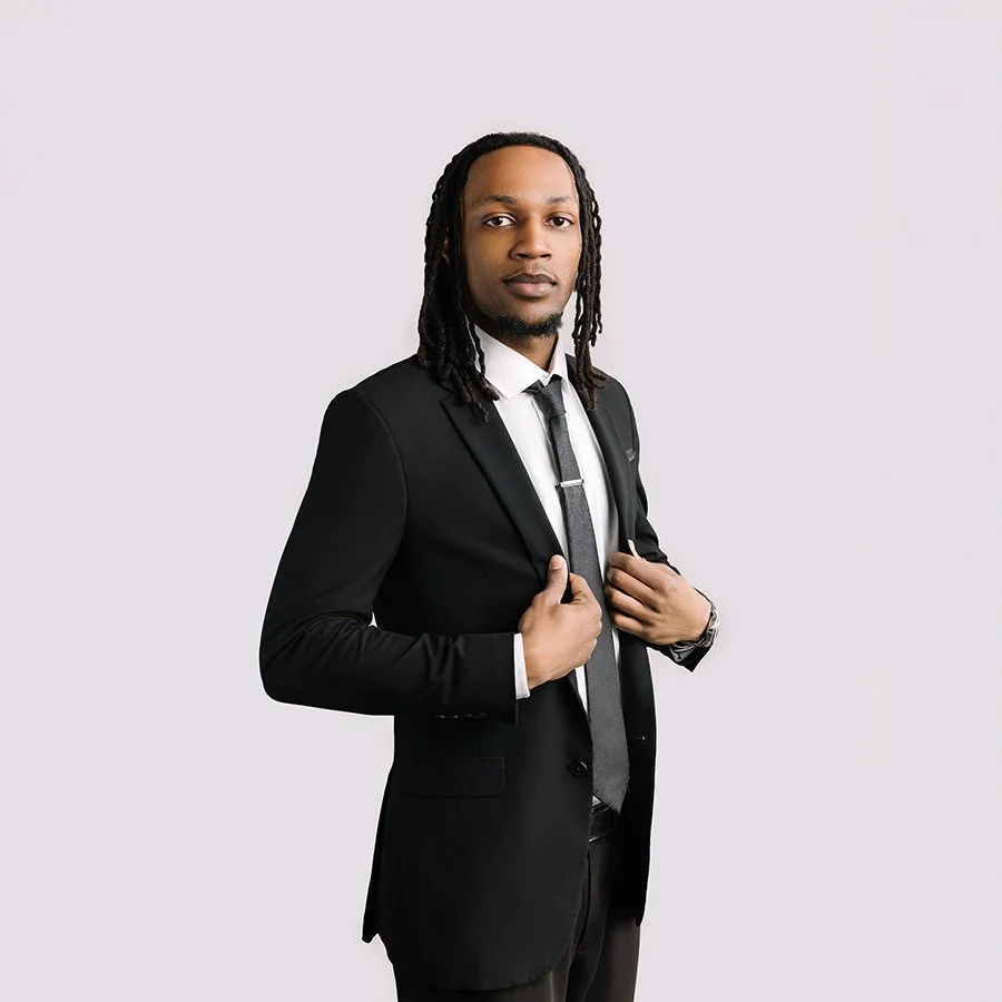 A man in a black suit, white shirt, and gray tie standing against a plain, light-colored background, adjusting his jacket.