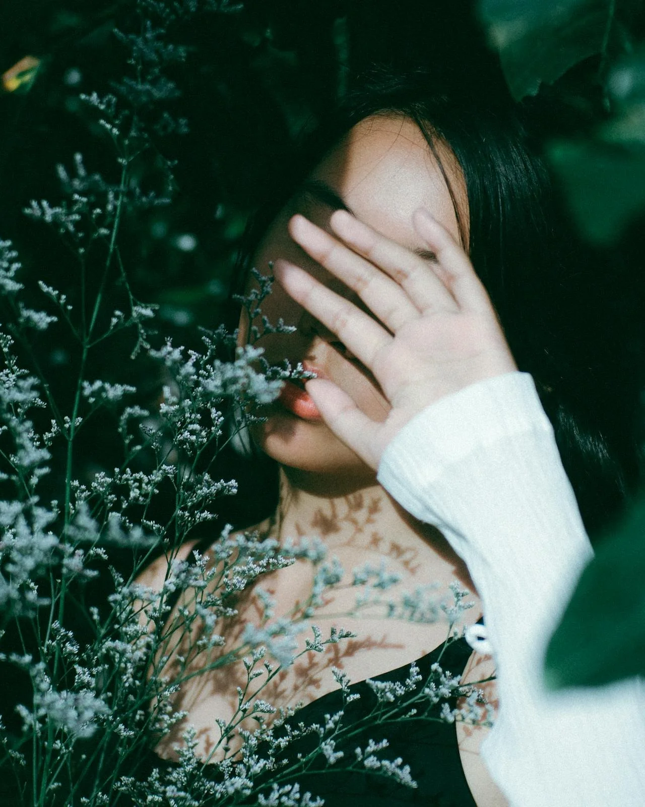 Asian woman covering her face with her hand among green plants, reflecting contemplation and cultural expectations