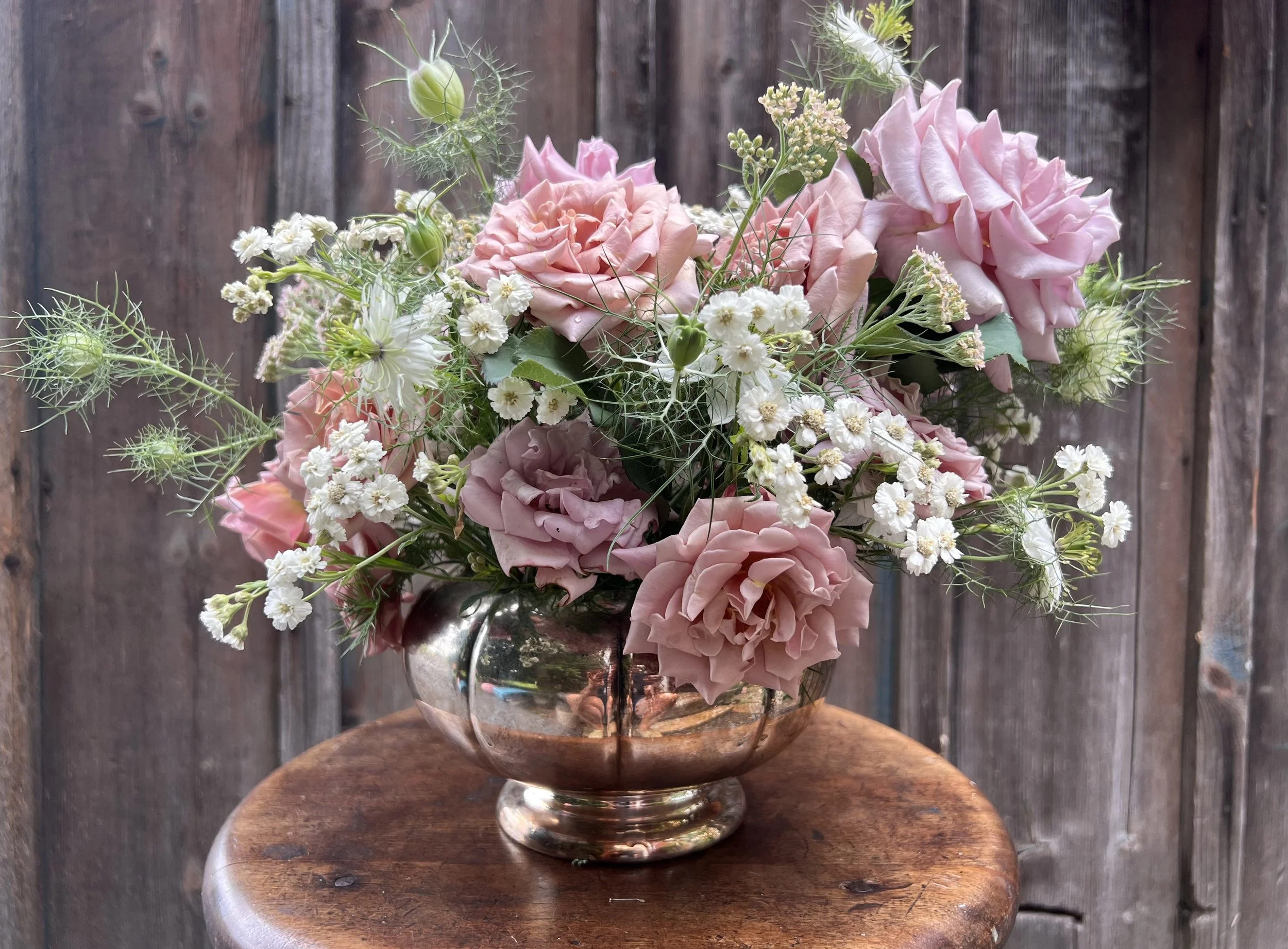A bouquet of pink and white roses, small white daisies, and green foliage in a shiny silver vase on a wooden table with a rustic wooden background.