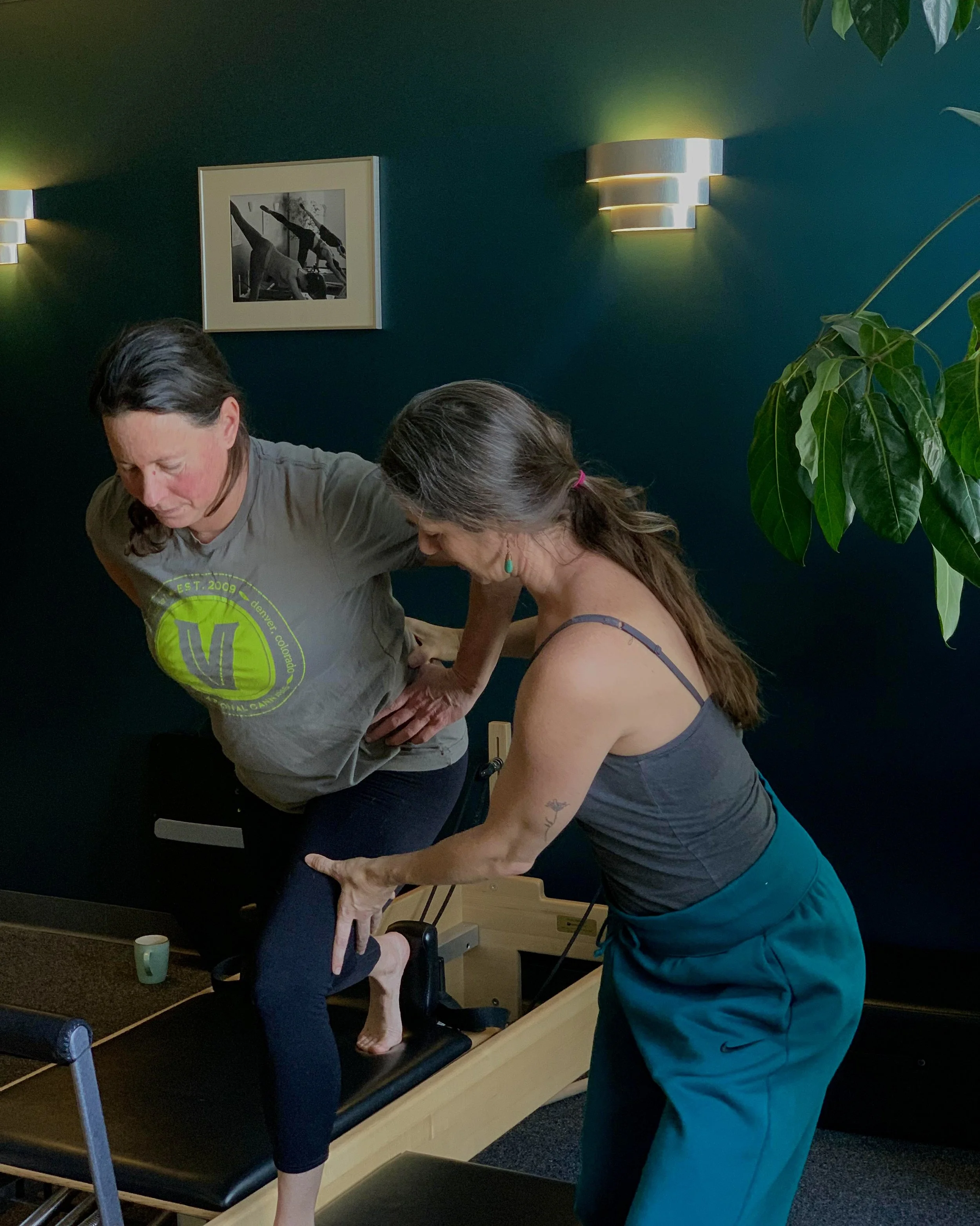 Two people practicing a balancing exercise on a Pilates reformer machine indoors.