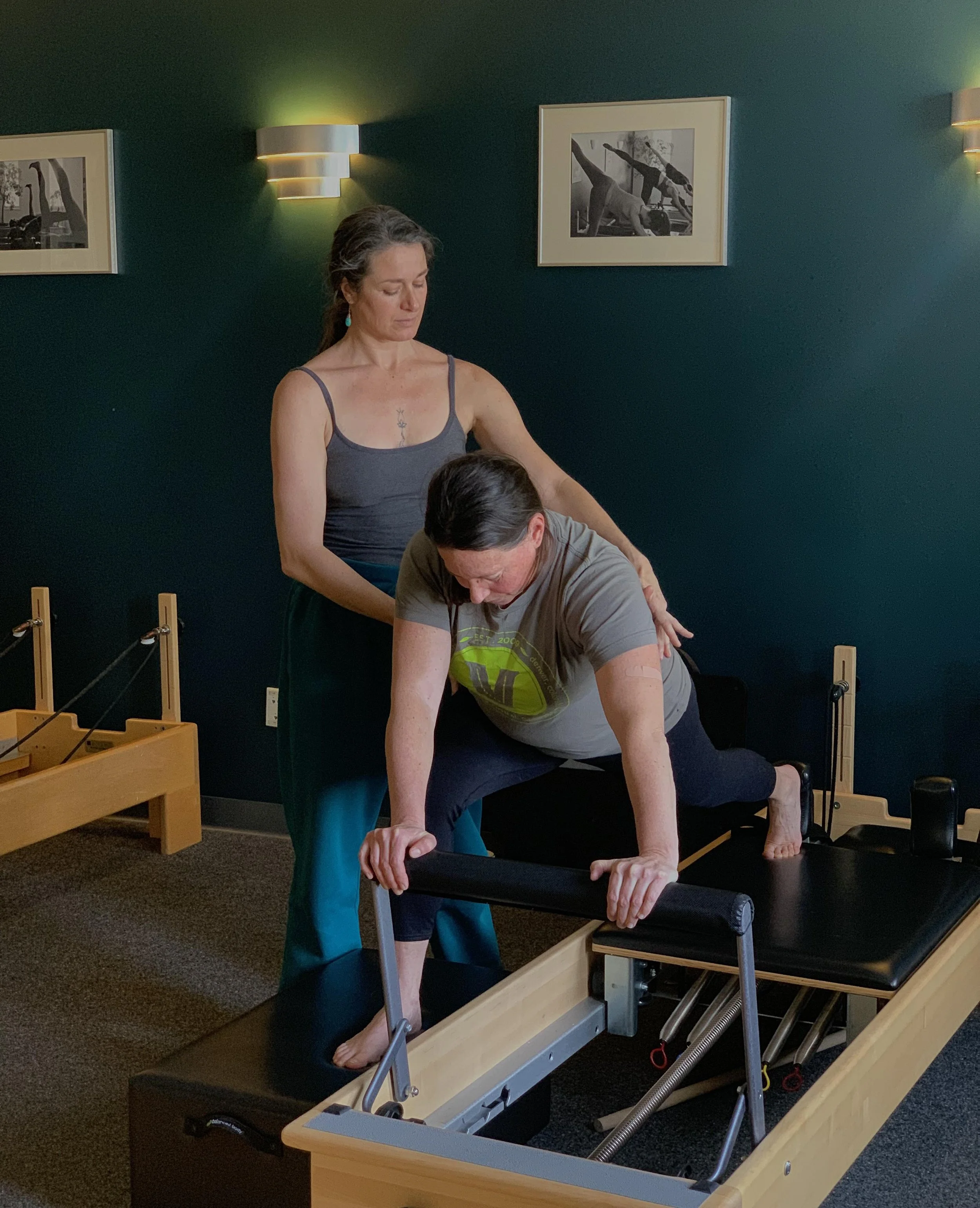 A Pilates instructor guiding a person on a reformer machine in a studio. The instructor's hands are on the person's back, assisting with posture and alignment. The room has dark walls and framed pictures. The person is kneeling on the reformer, holding the bar.