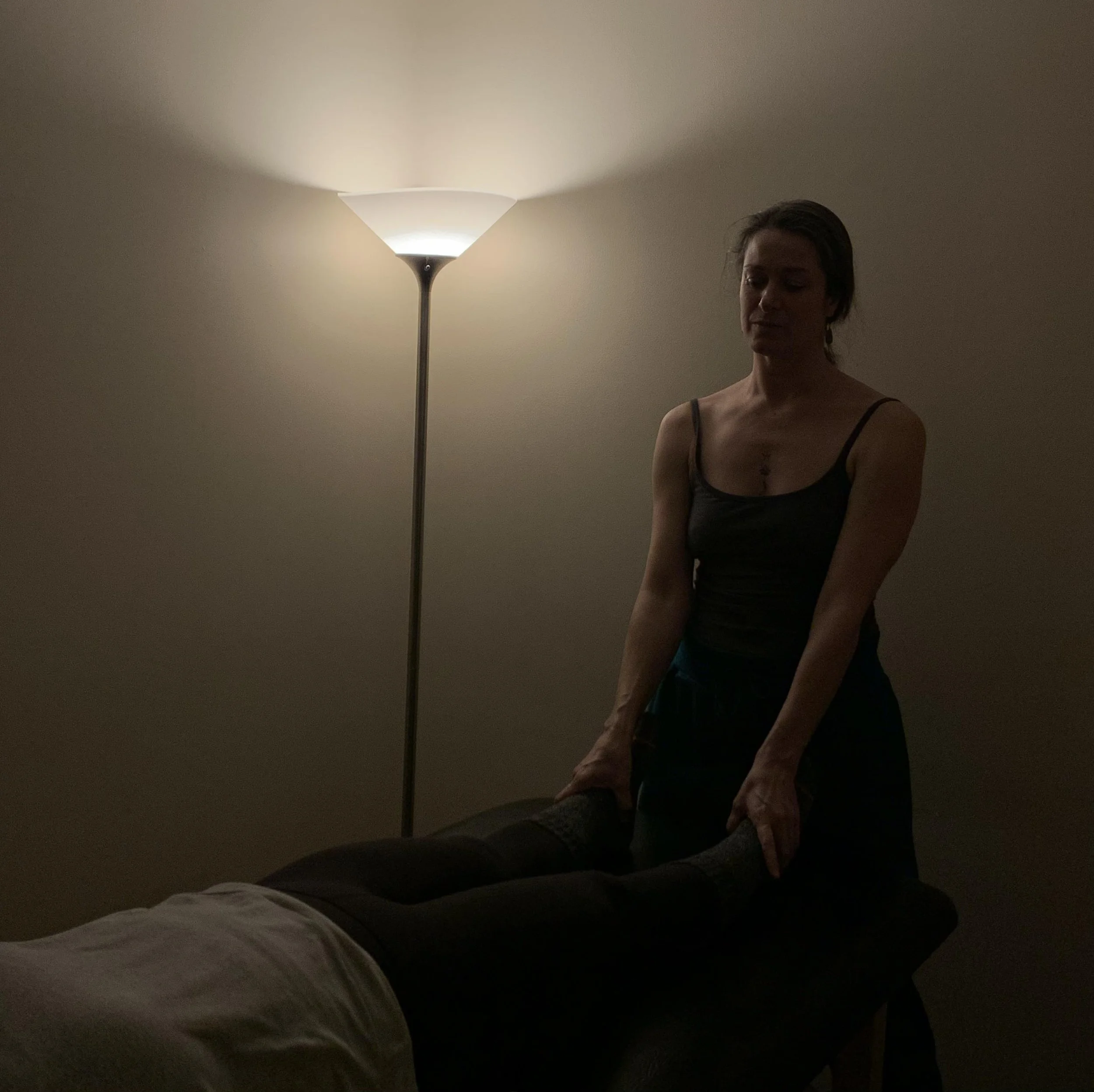 A dimly lit room with a woman practicing craniosacral therapy on a person lying on a table, illuminated by a floor lamp.