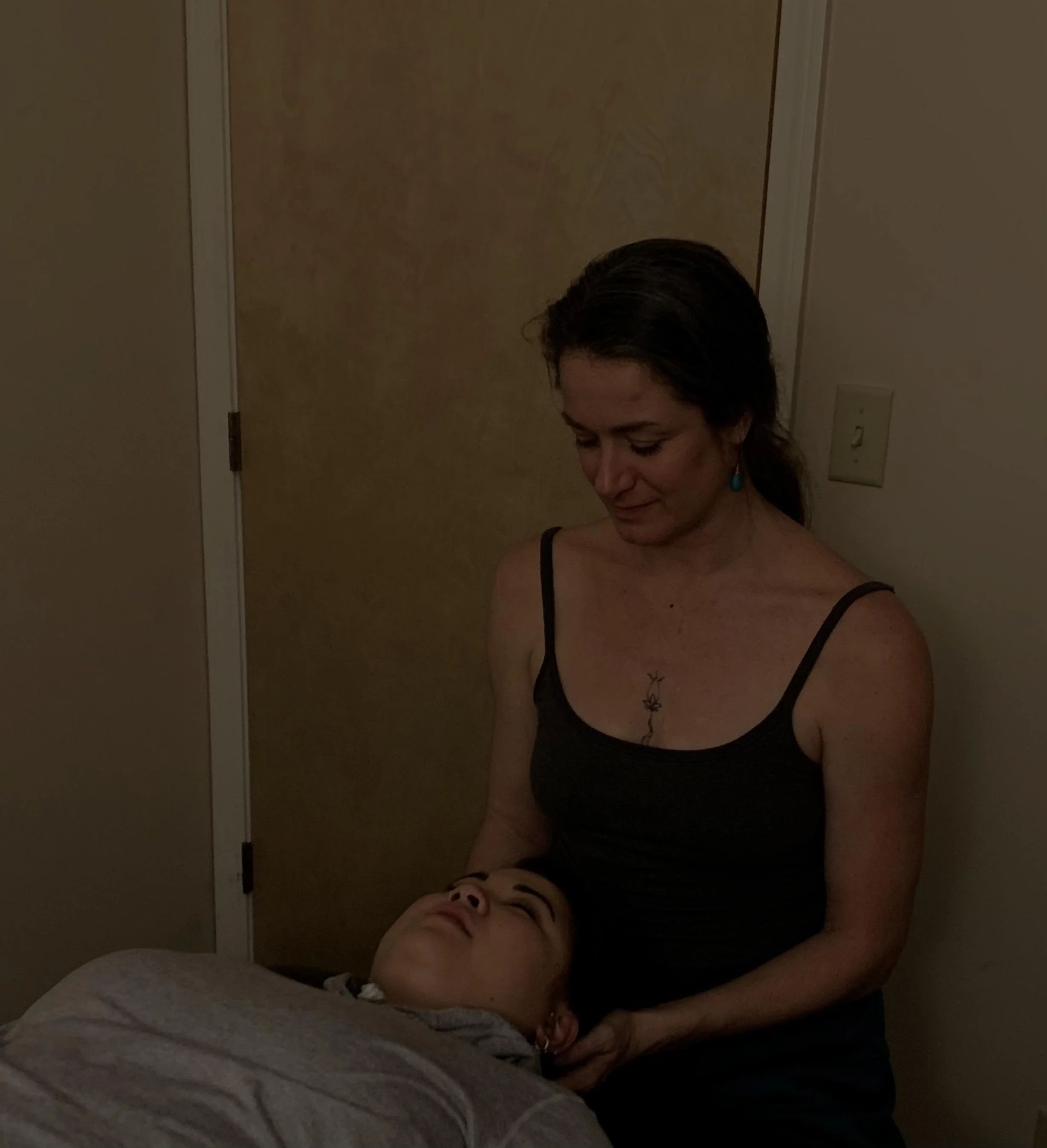 A woman giving craniosacral therapy to a person lying down in a dimly lit room.