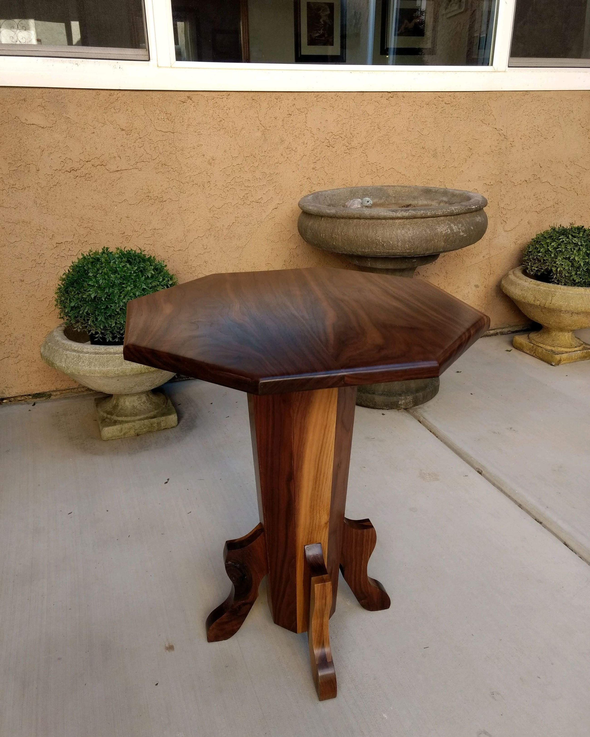 A wooden table with a curved edge, standing on a concrete patio, with two stone planters containing green bushes and a wall with a window in the background.