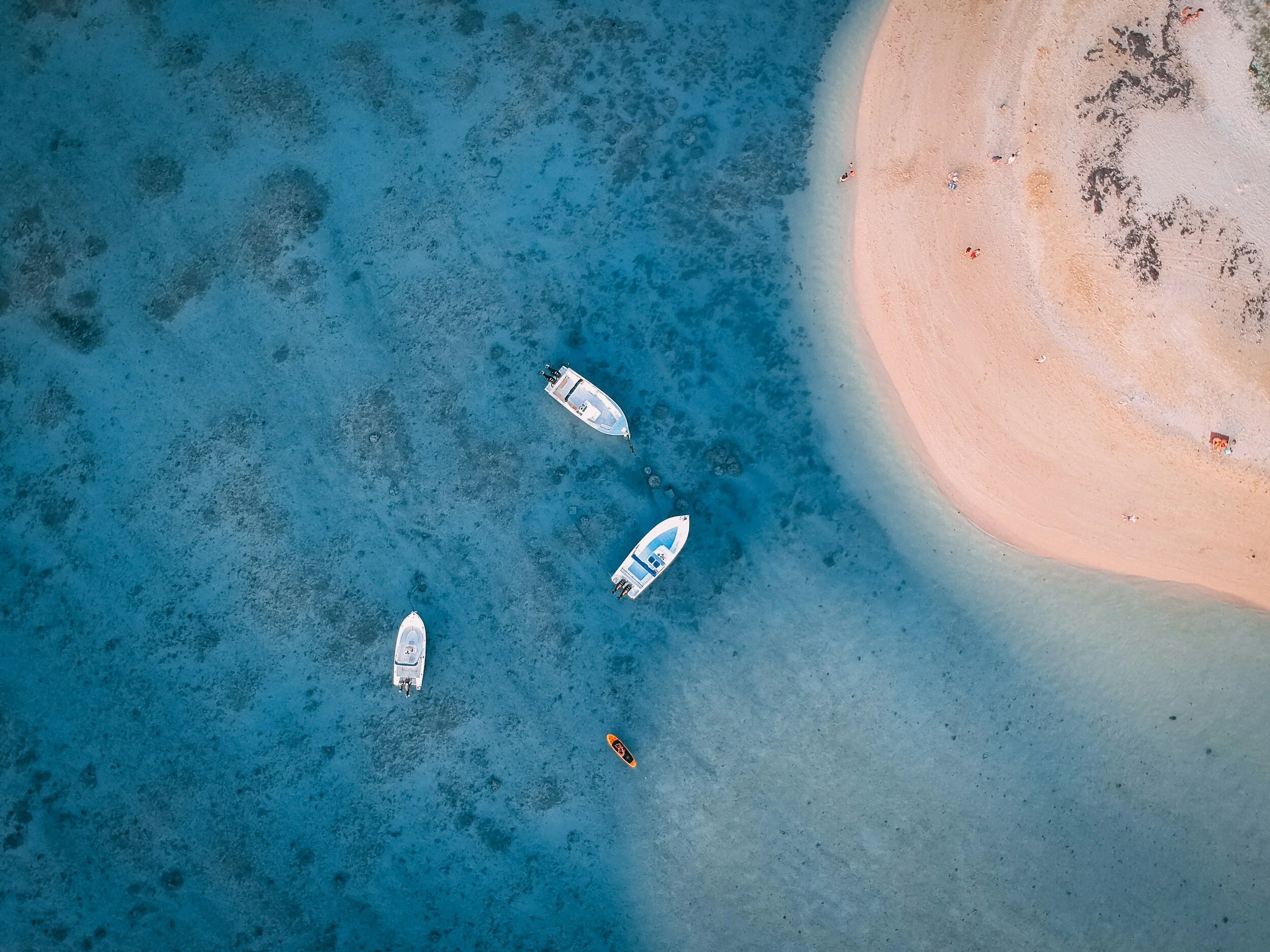 Aerial view of a beach showing three boats floating on the water near the shore, with a sandy beach and some people and objects visible on the sand.