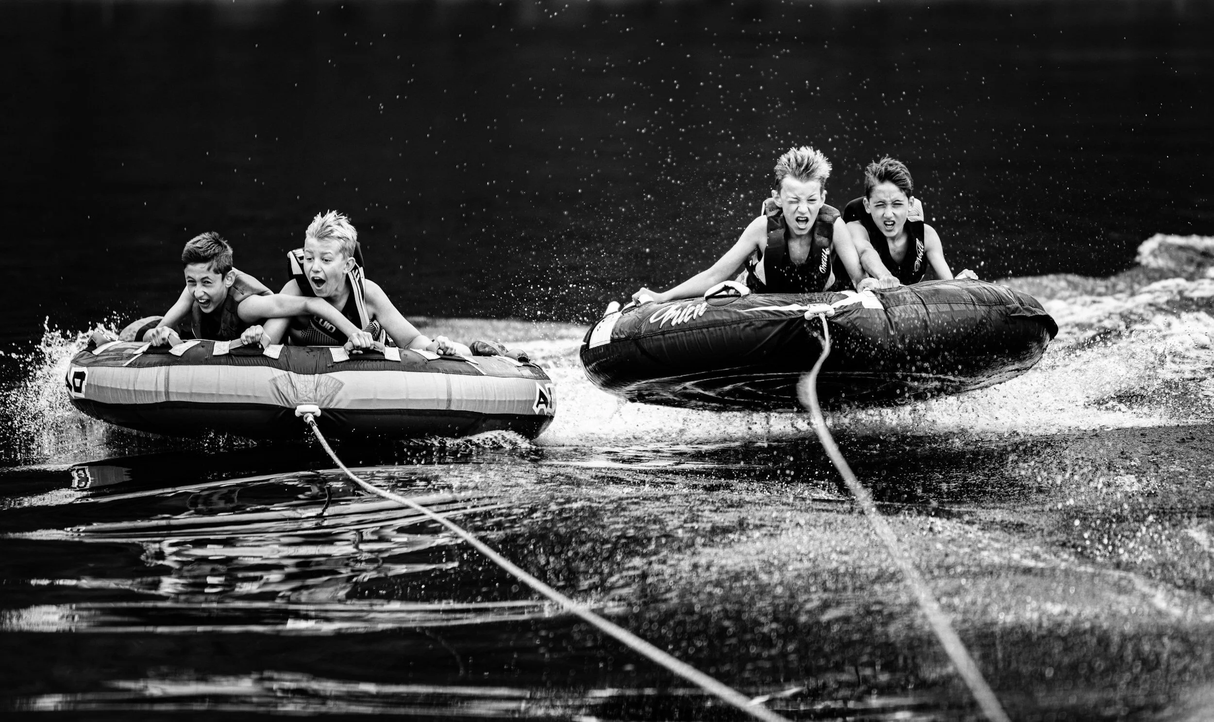 Four children riding inner tubes pulled by a boat on a lake, all showing expressions of excitement and determination, in black and white.