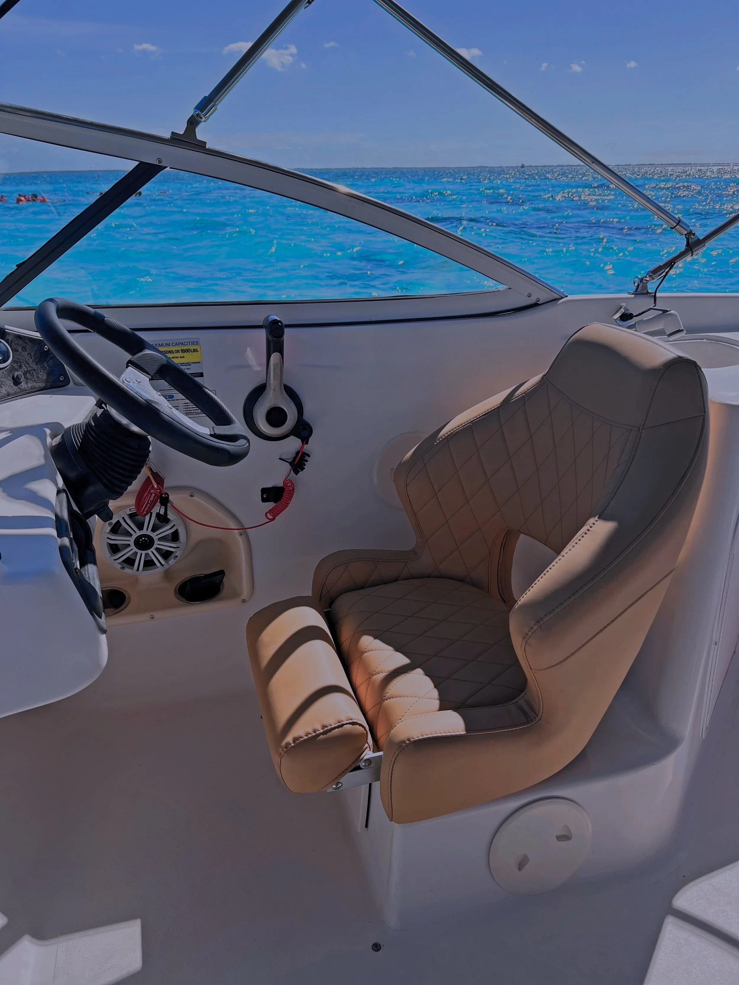 Interior view of a boat cockpit showing a beige captain's chair, steering wheel, control lever, and a speaker, with blue water and sky visible through the windshield.