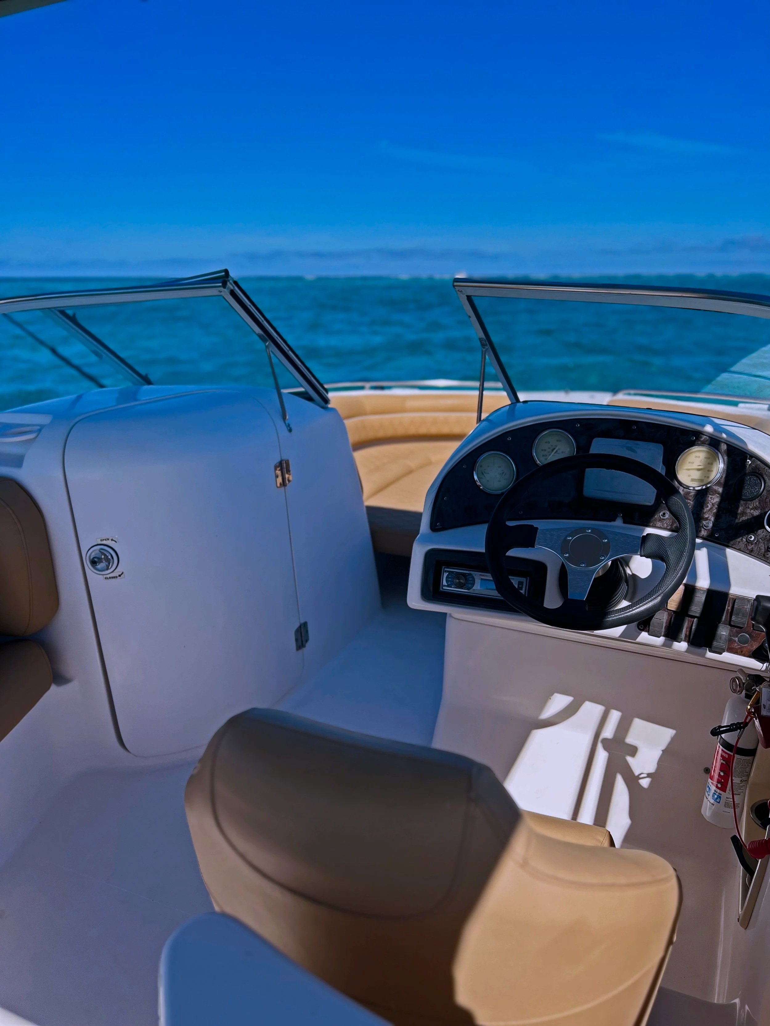 View of the interior of a boat cockpit with a steering wheel, dashboard gauges, and seating, overlooking a blue ocean and sky.