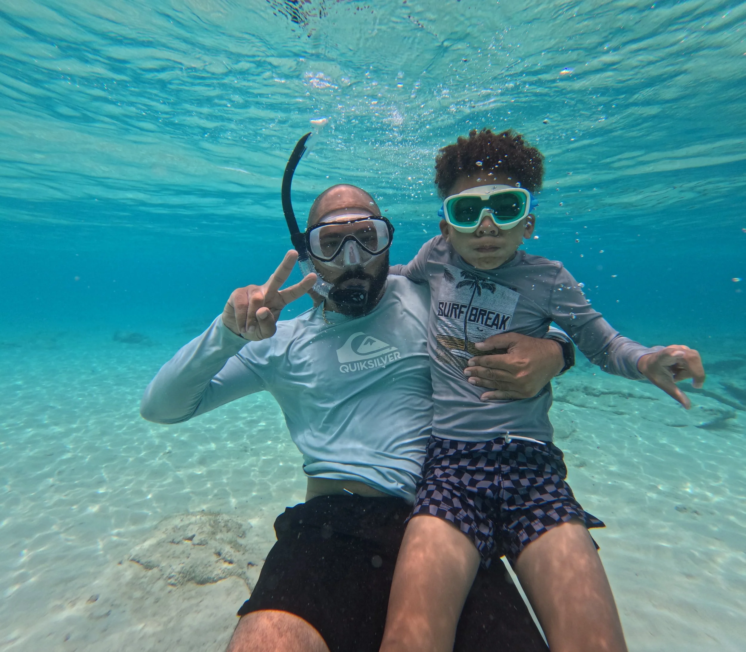 A man and a boy in snorkeling gear underwater, posing for a photo. The man is making a peace sign with his fingers. Both are wearing swimshirts and goggles.
