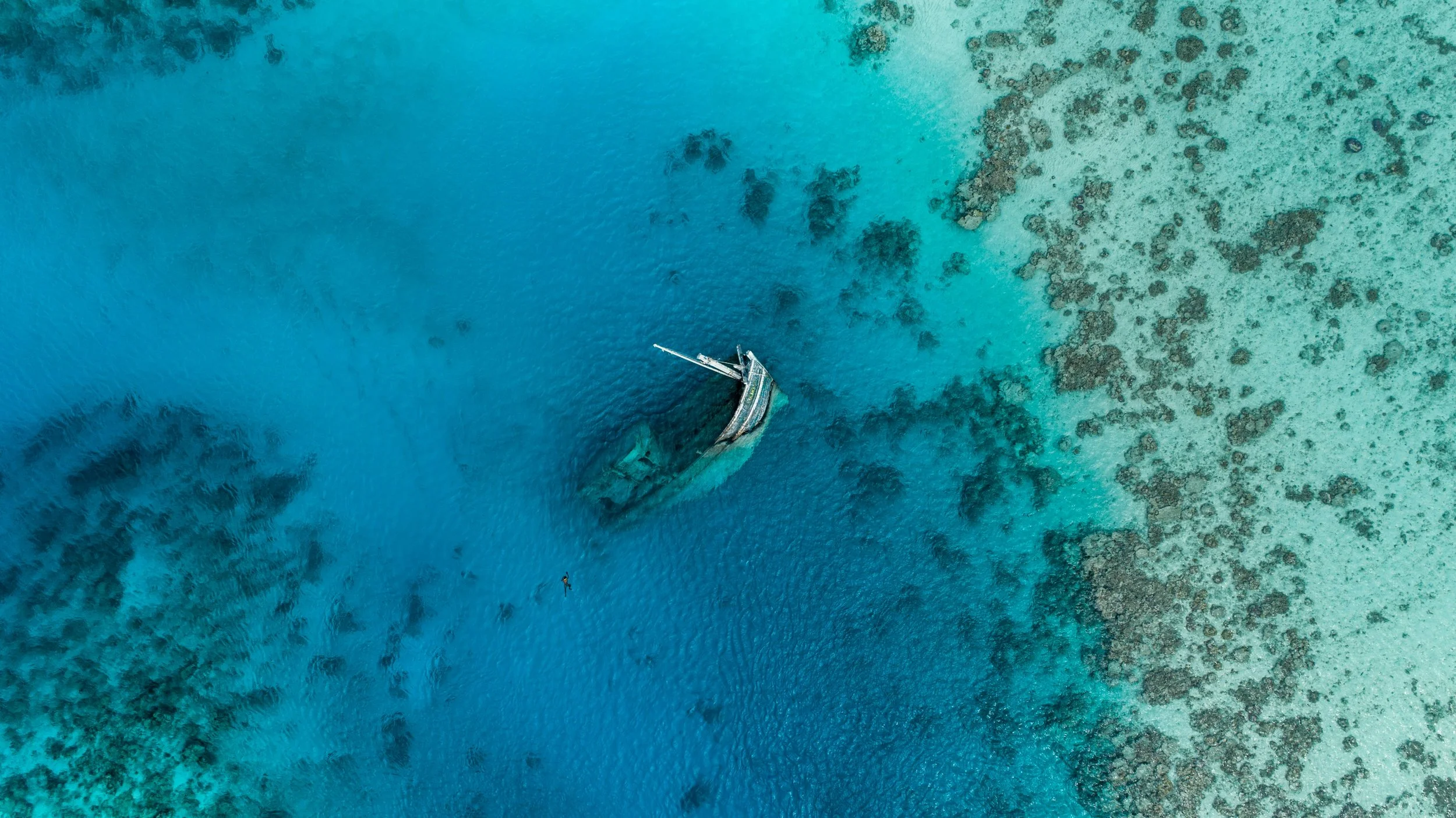 A sunken boatwreck is visible in shallow, turquoise waters with coral reefs around it, captured from an aerial view.