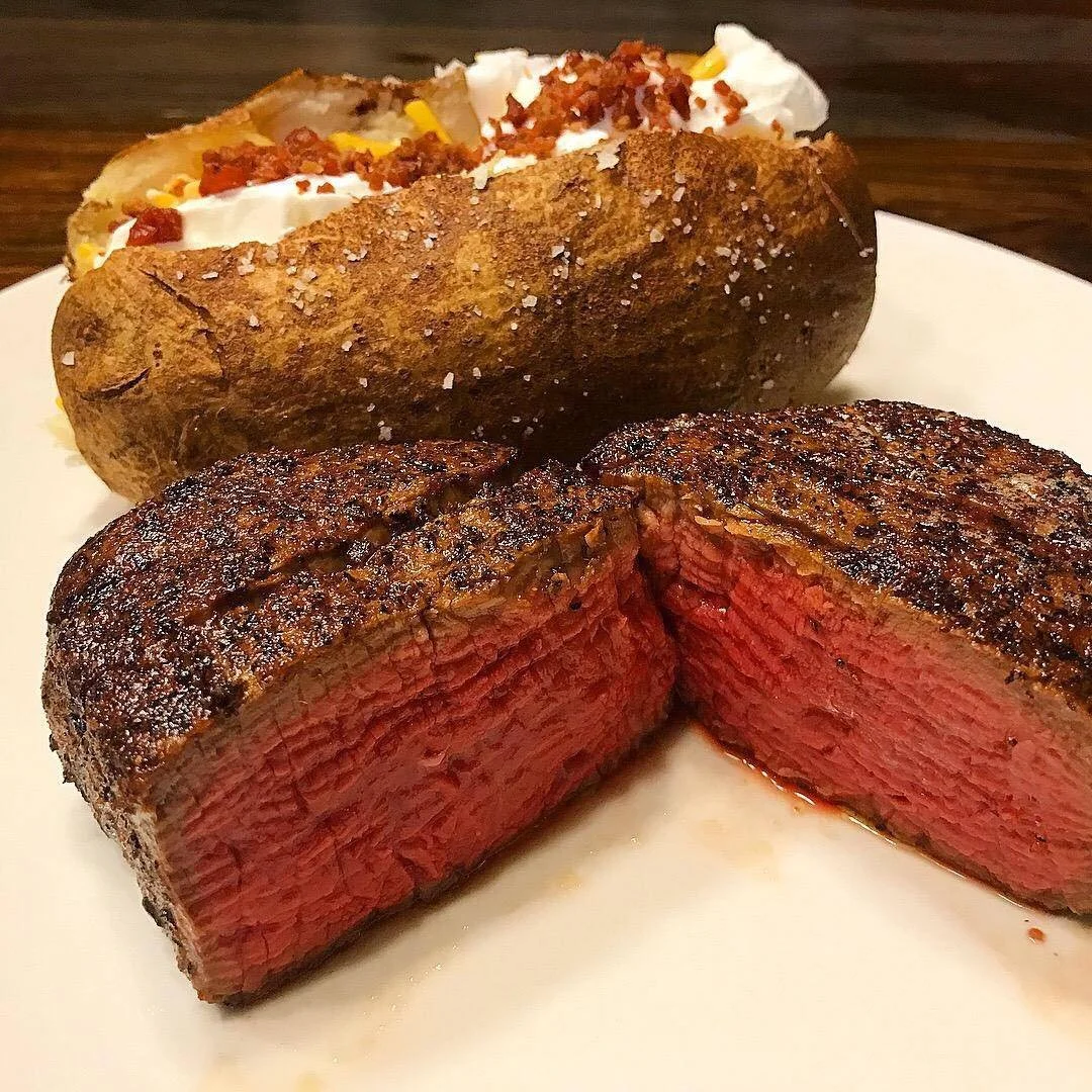 A cooked steak cut in half revealing a pink center, served on a white plate along with a baked potato topped with sour cream, shredded cheese, bacon bits, and chives.