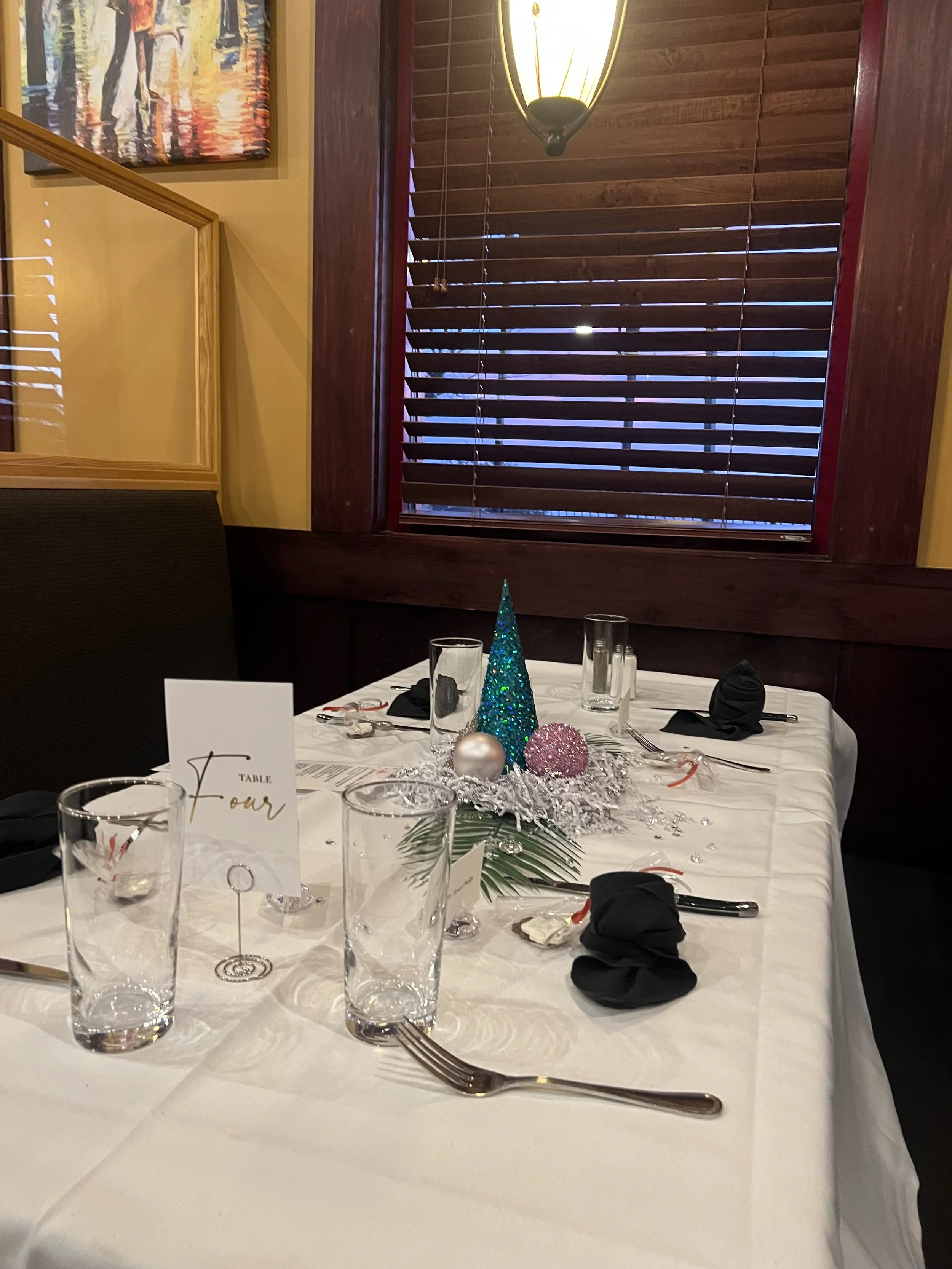 A decorated table set for a celebration with a small Christmas centerpiece. The centerpiece includes a teal glittery cone, a pink textured ball, a silver ball, and sprigs of greenery and silver tinsel. The table has black cloth napkins, silverware, and empty glasses. A table card reads 'Table Four.' The background features wooden blinds and a wooden-paneled wall with a hanging light fixture.