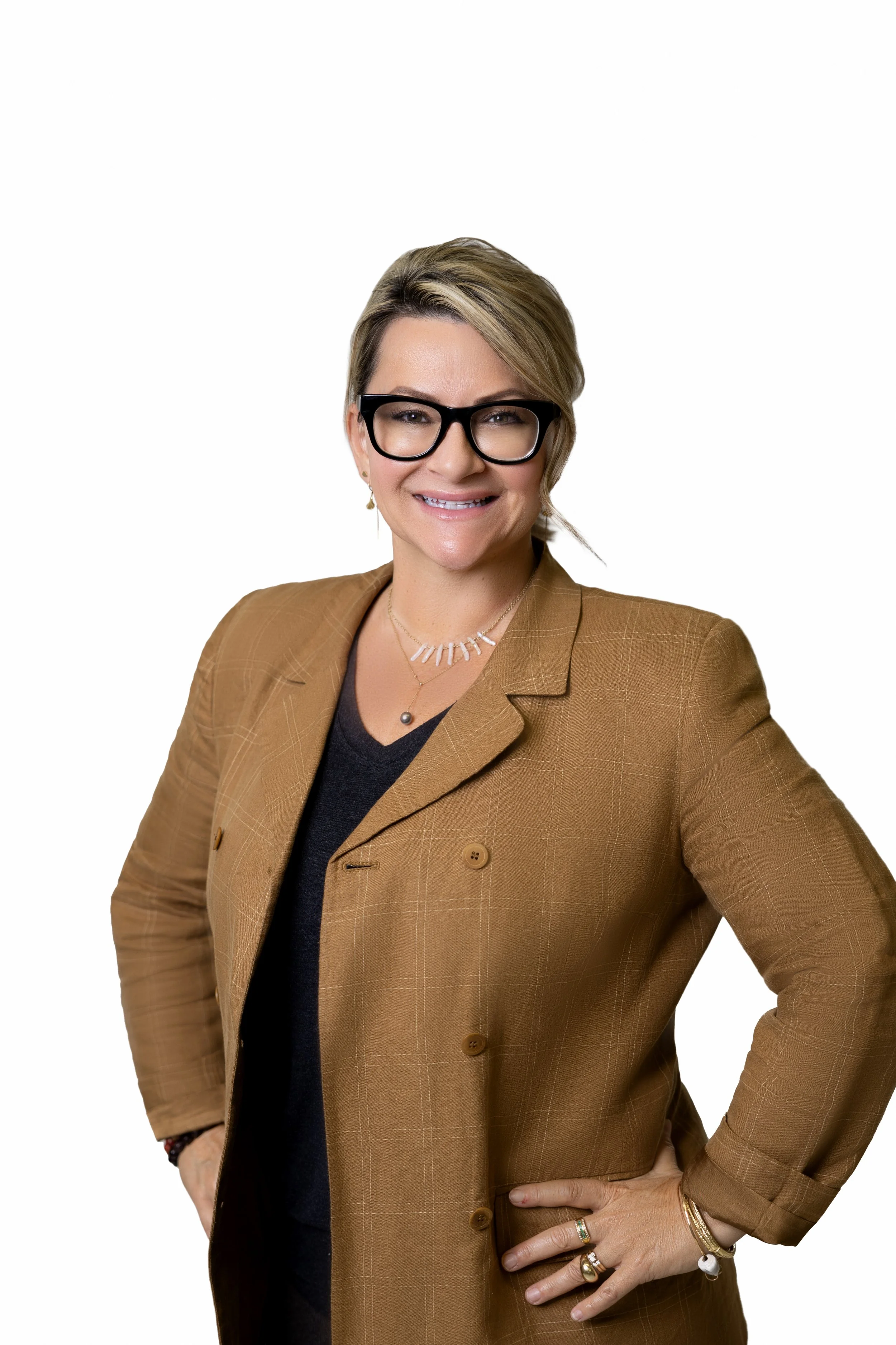 A woman with short blonde hair, black glasses, wearing a brown blazer and a black top, smiling with her hand on her hip, accessorized with rings, bracelets, and jewelry, against a white background.