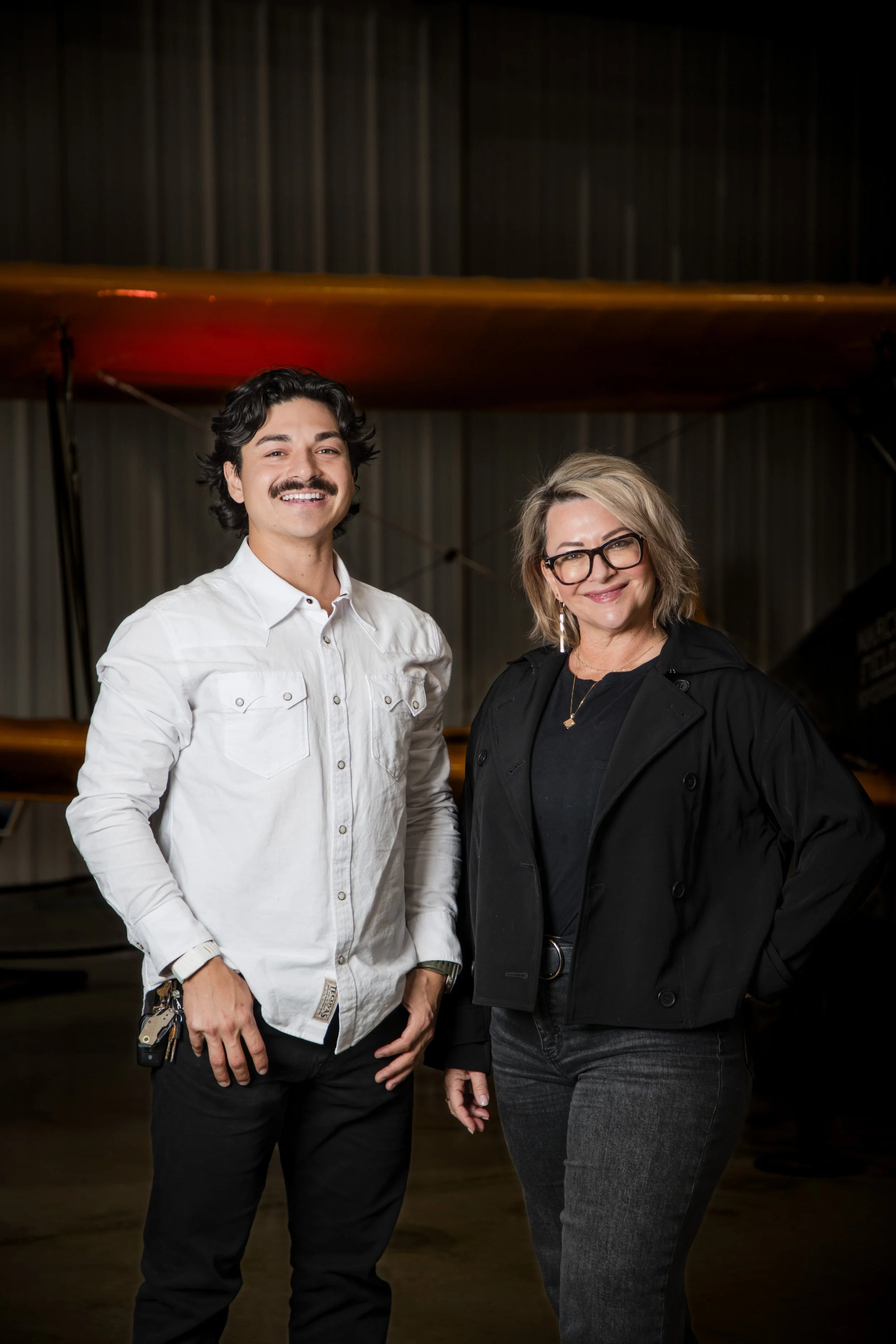 A man with dark wavy hair, a mustache, and a white shirt, standing next to a woman with blonde hair, glasses, and a black jacket, in front of a dark curtain and a wooden backdrop.