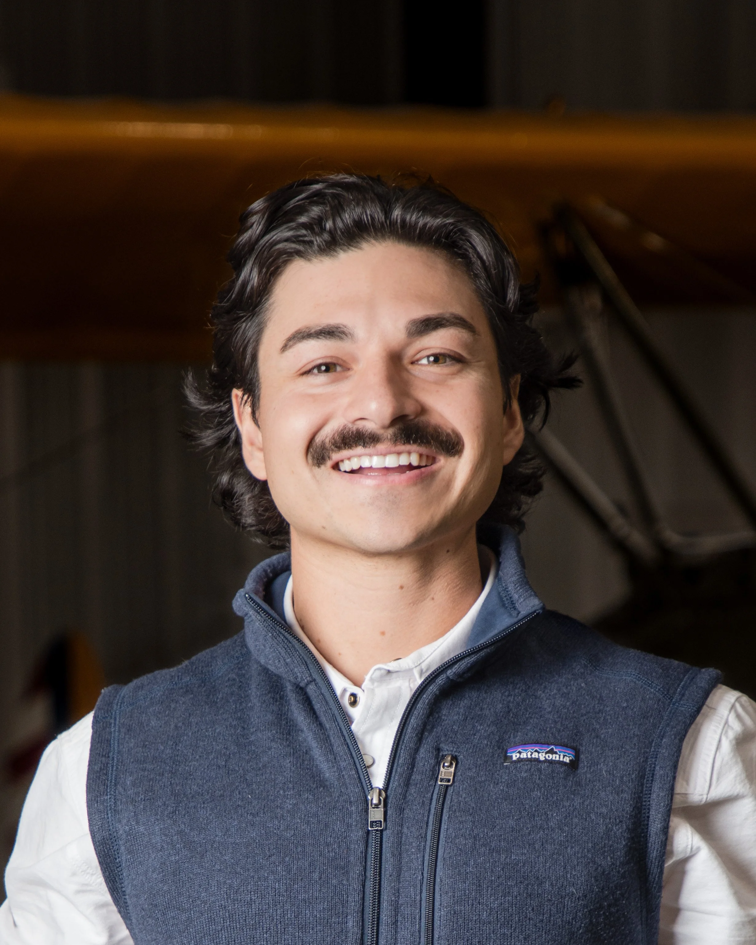 Close-up of a smiling man with dark, wavy hair, wearing a white shirt and a navy blue Patagonia vest, indoors with blurred background.