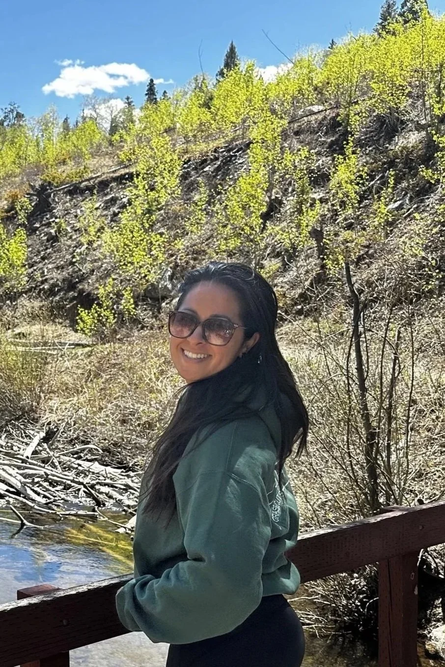 A smiling woman in outdoor gear, including a hat, hoodie, and backpack, standing on a wooden bridge with a mountain and forest in the background.