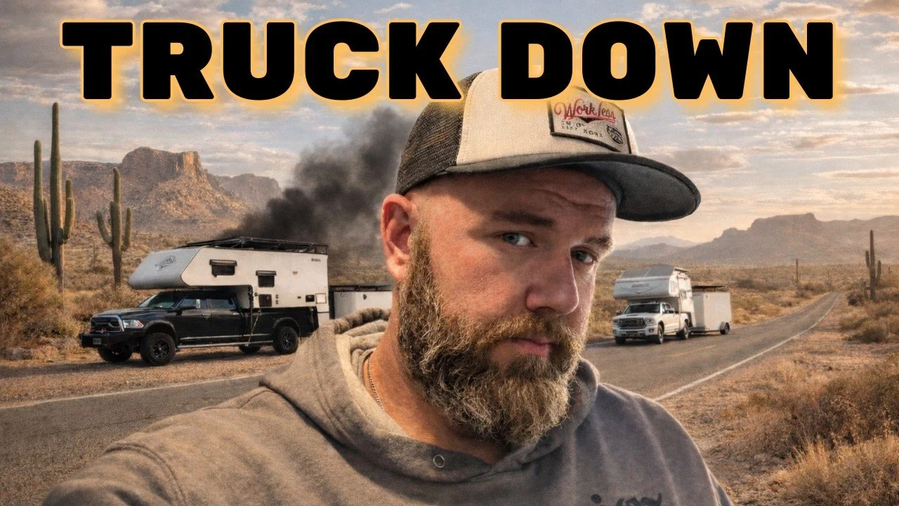 A man with a beard wearing a baseball cap and hoodie taking a selfie on a desert highway. In the background, two trucks are stopped, with one of them emitting black smoke. The landscape includes cacti and mountains under a cloudy sky, and the text 'T