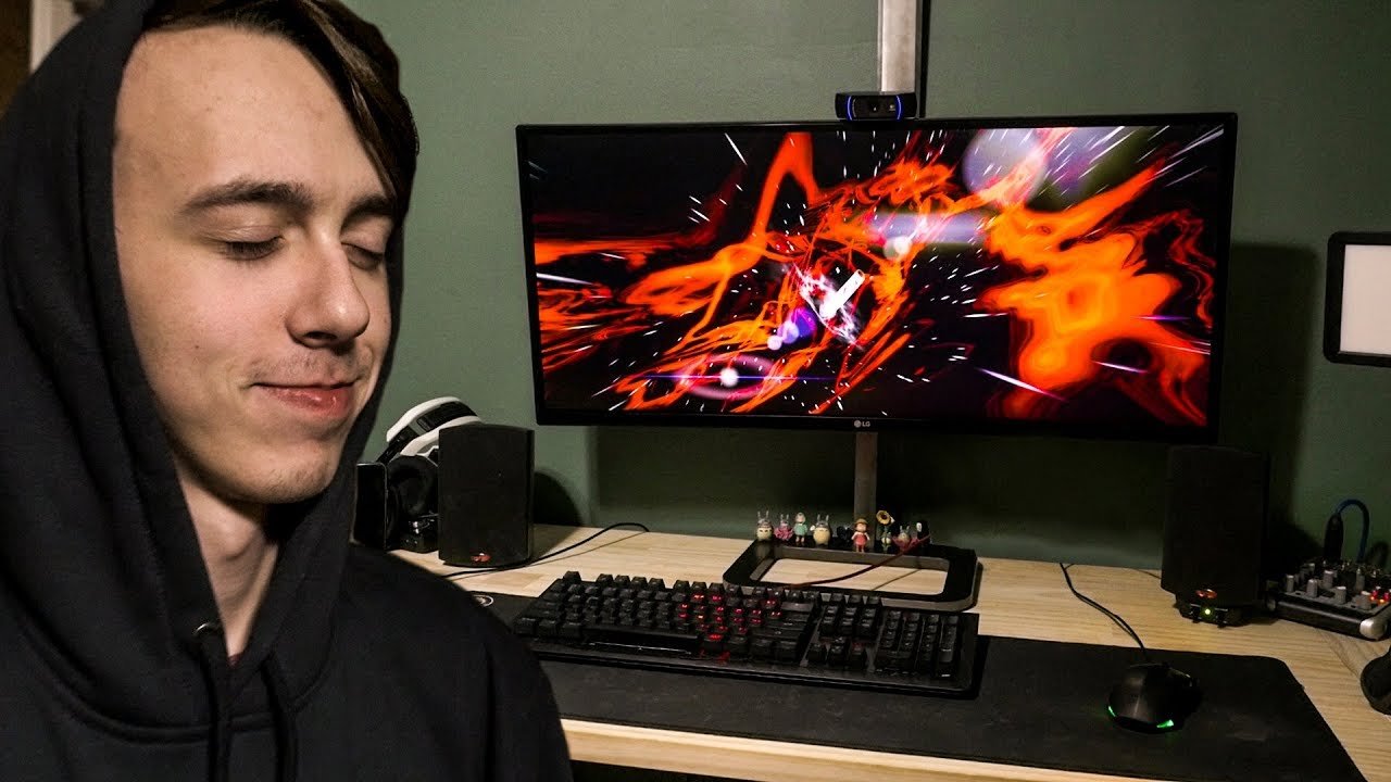 A person with closed eyes smiling in front of a computer setup with a curved monitor displaying an abstract, colorful digital design in red, orange, purple, and black, with speakers and a keyboard on the desk.