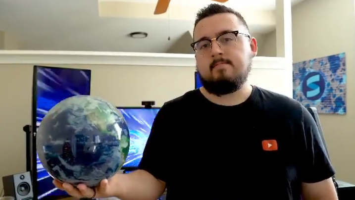 Matt Philie holding a globe in his right hand, standing in front of a desk with multiple computer monitors in a room with white walls and a ceiling fan.