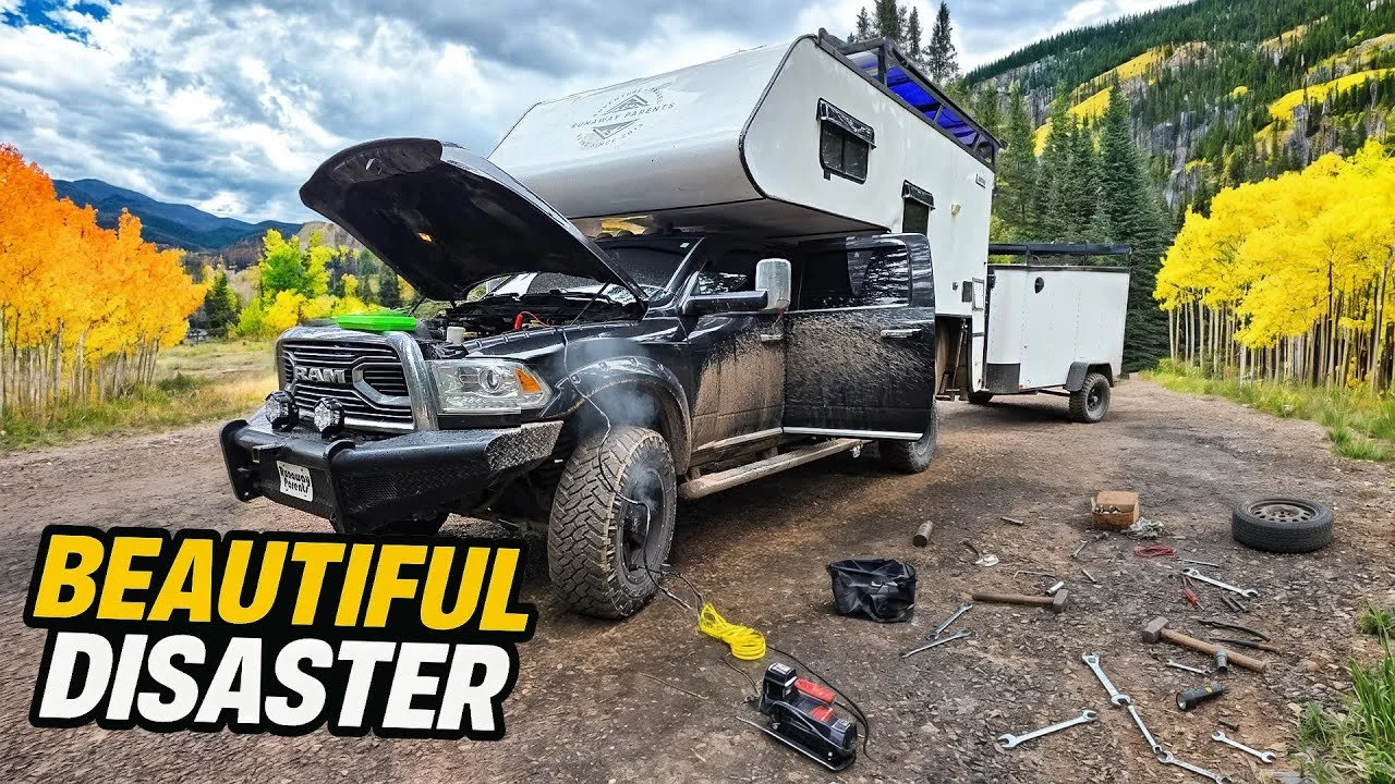A black pickup truck with its hood open and a trailer hitched to it, parked on a dirt road surrounded by colorful autumn trees and mountains in the background. Tools and spare tire are scattered on the ground.