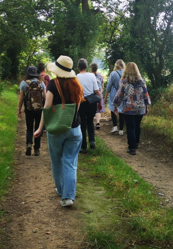 A group of different people are walking along a country lane on a summer's day. In the distance a field is bathed in sunlight, and is framed by green trees.
