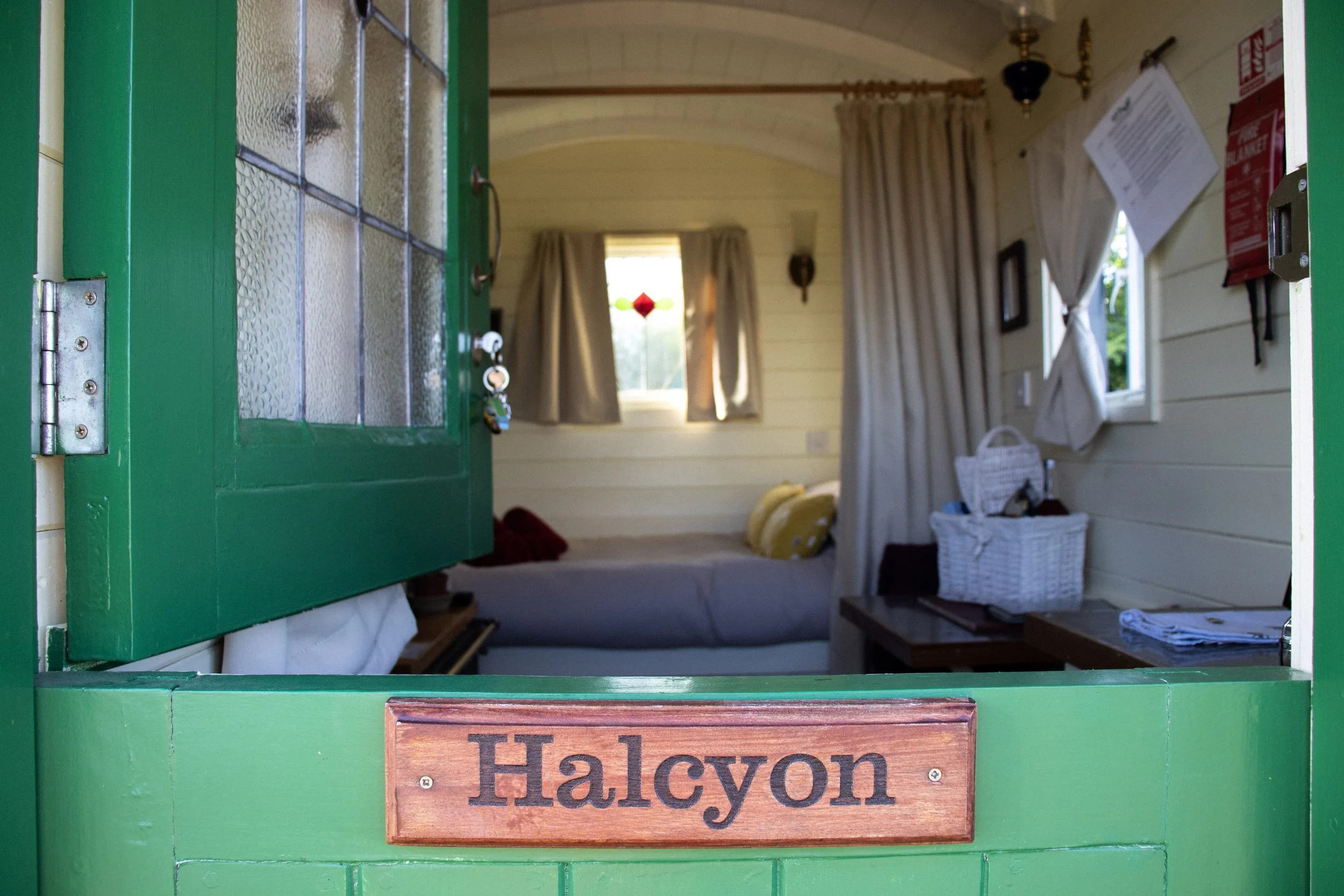 A green half door is open at the top, revealing a cosy shepherd's hut inside with a bed, curtains and basket of gifts. On the closed bottom half is a wooden sign reading 'Halcyon'