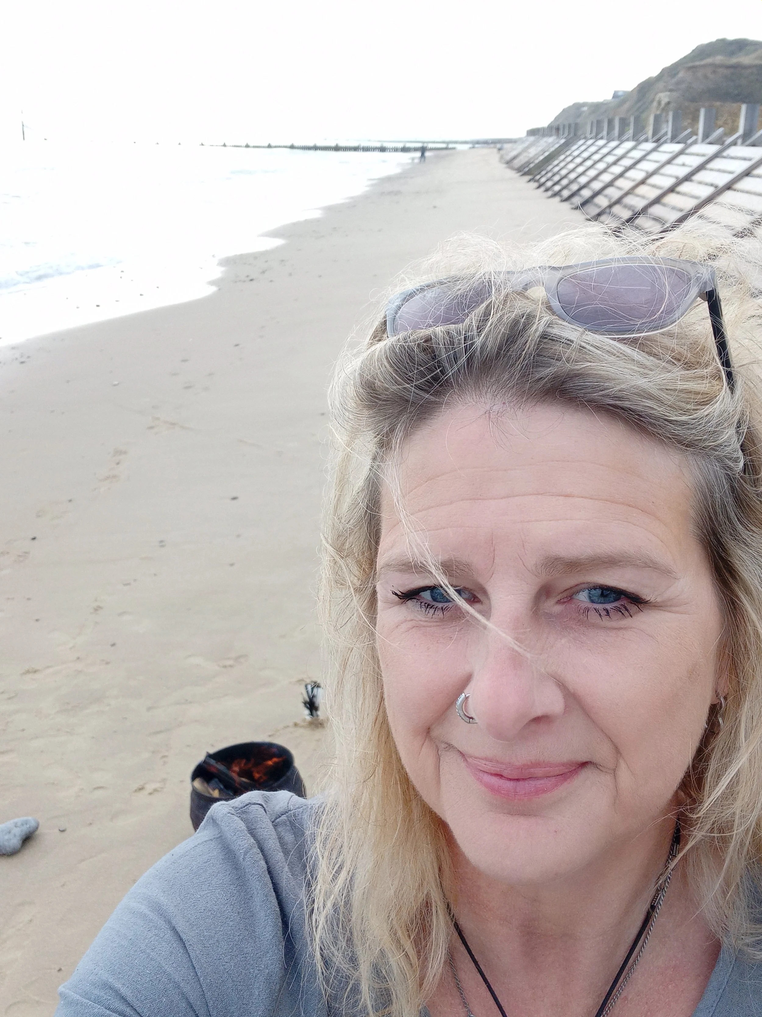 Sue is looking at the camera, with windswept blonde hair and sunglasses resting on top of her head. She is on a beach with the sea crashing against the sand nearby