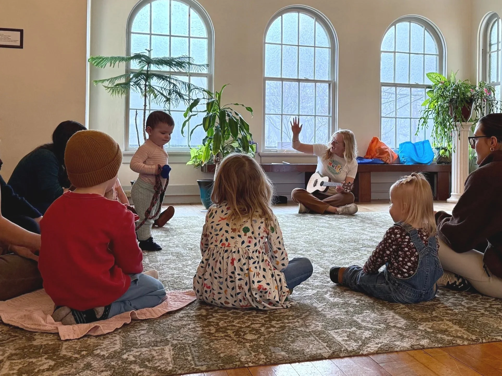 A woman with a guitar sitting on the floor near a large window in a room, engaging with a group of children and adults during a musical activity.