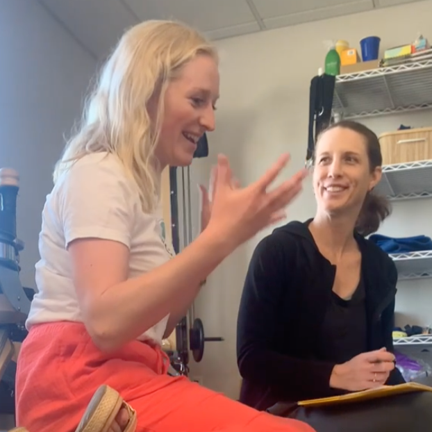 Two women sitting and talking in a room with shelves holding various items in the background.