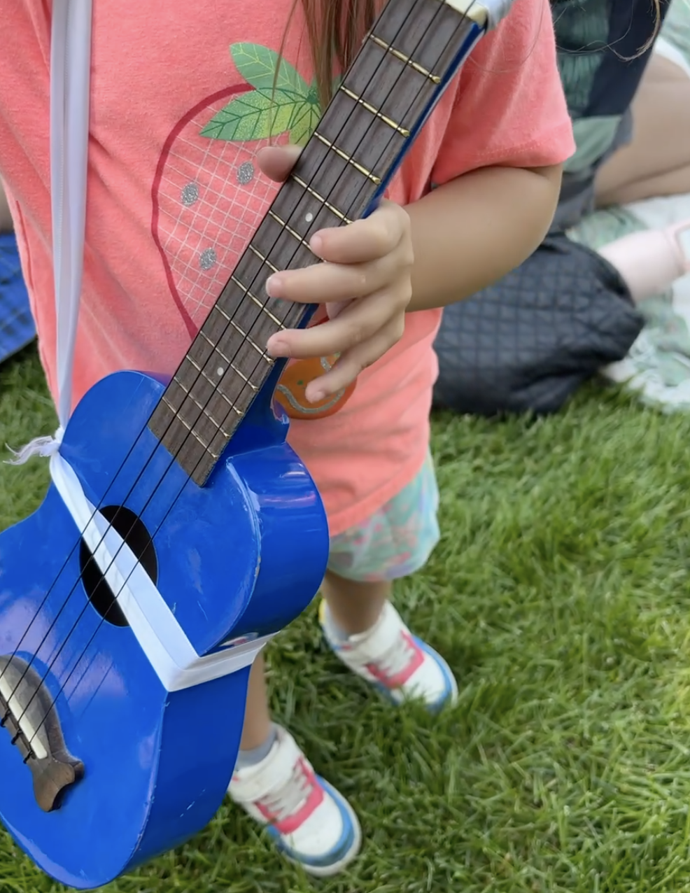 Child holding a small blue toy guitar outdoors on grass, wearing a pink shirt with an apple graphic and colorful shorts, with people and objects in the background.