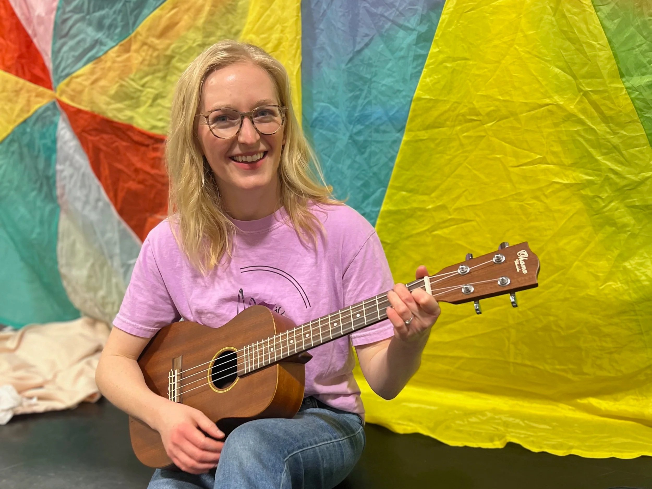 A smiling woman with glasses and blonde hair playing a ukulele in front of a colorful, crinkled paper background.