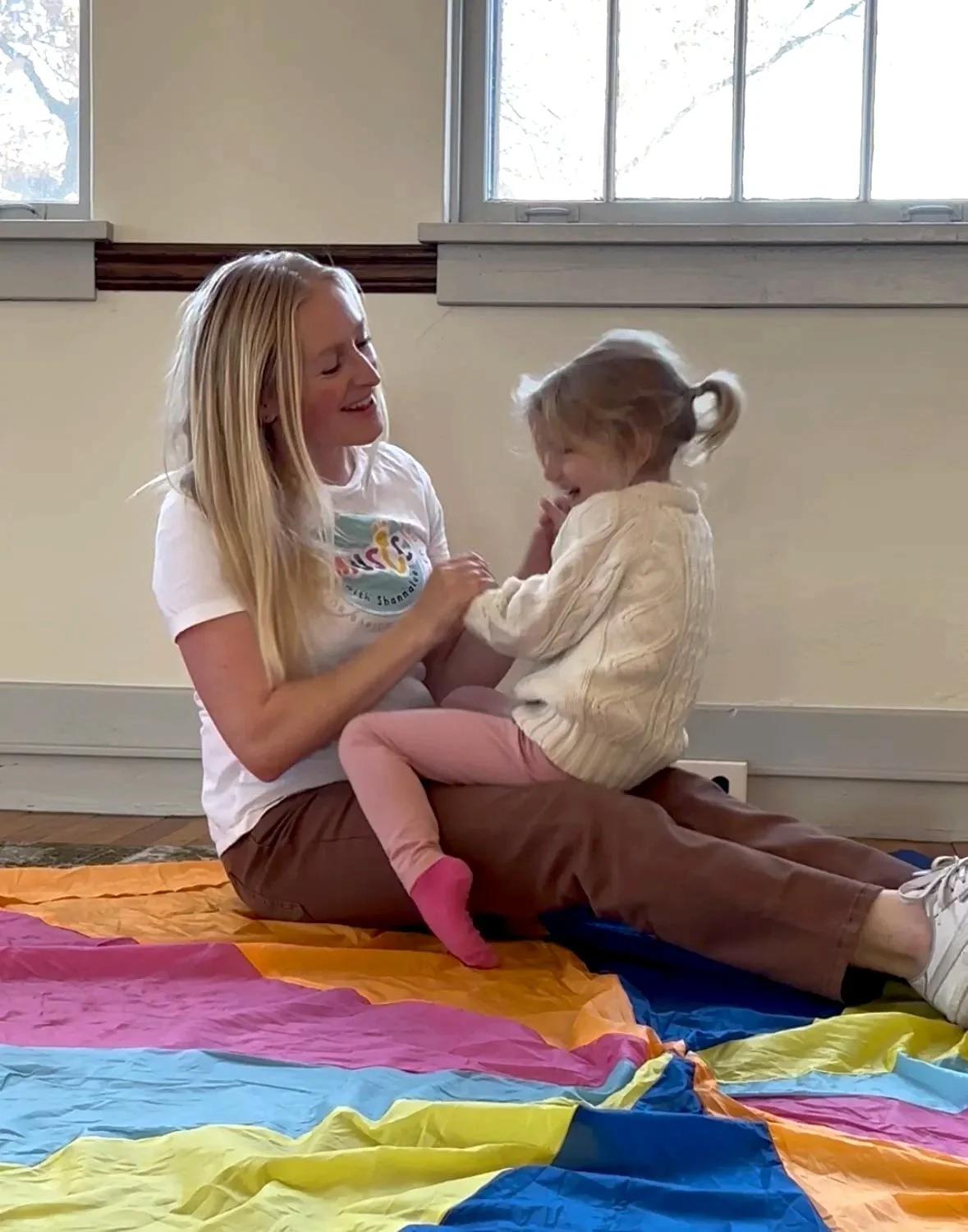 A woman and a young girl playing and laughing on a colorful blanket indoors, near windows.