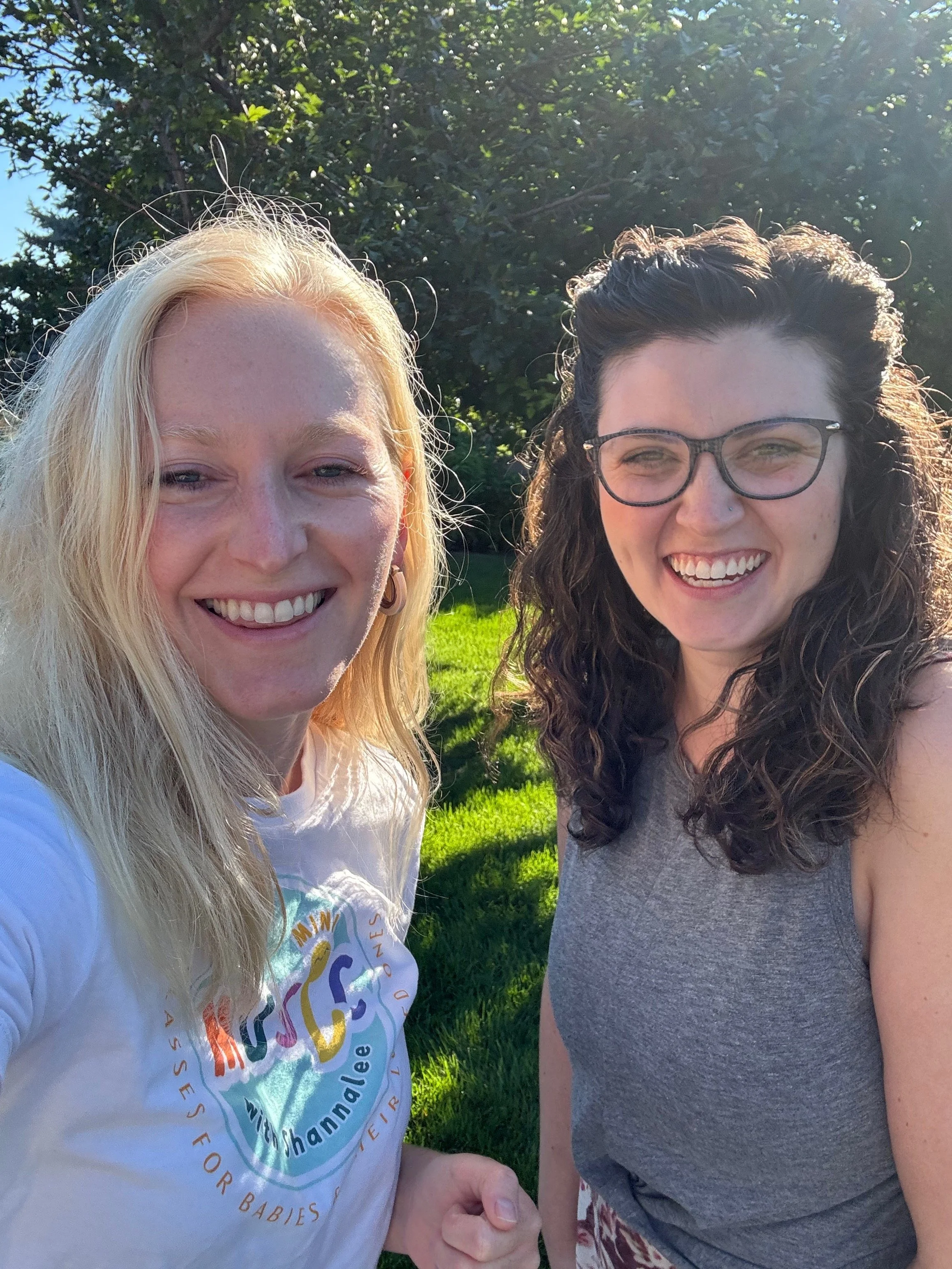 Two women smiling outdoors on a sunny day, standing on green grass with trees in the background.