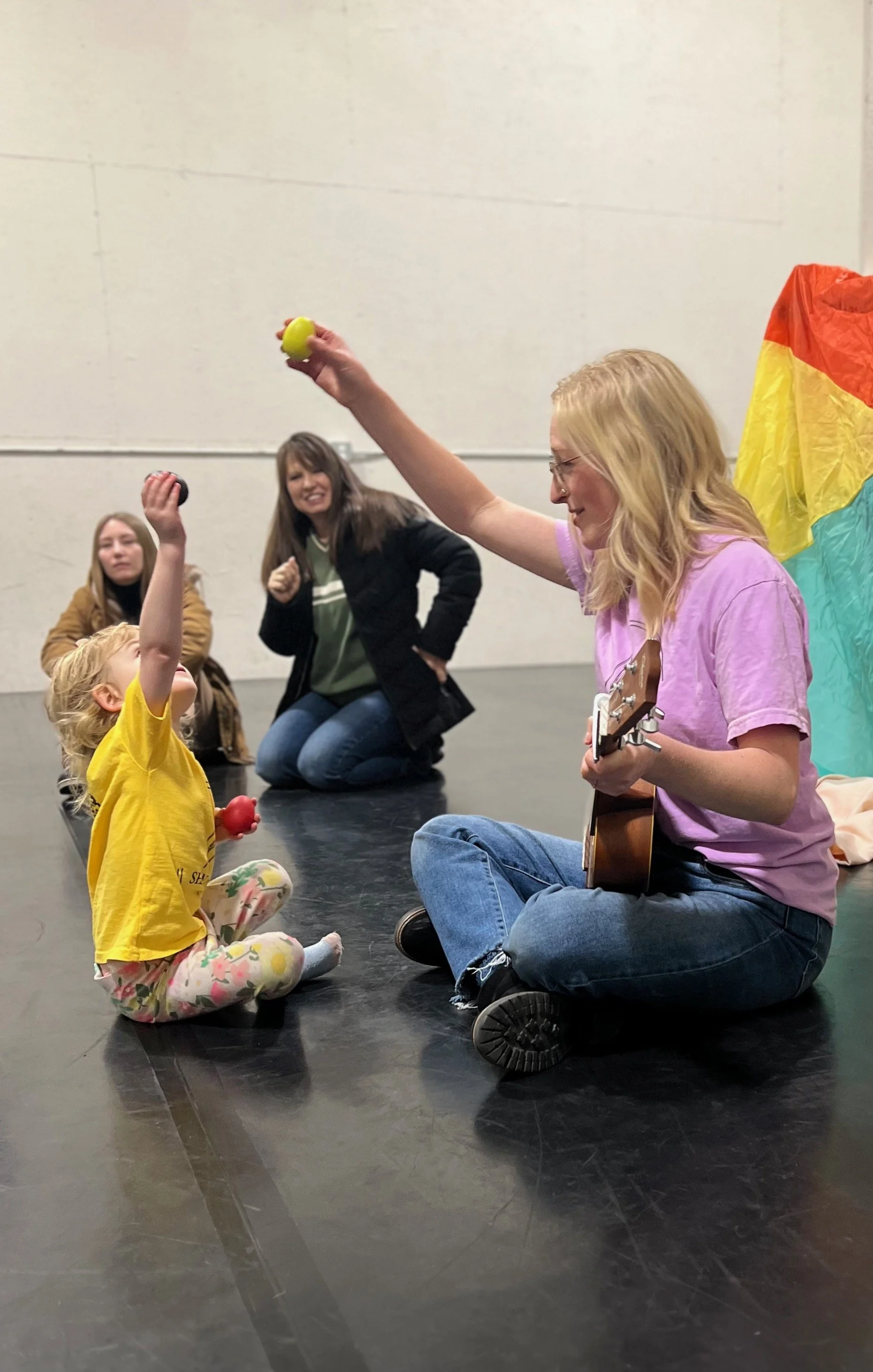 A woman playing a guitar sitting on the floor, giving colorful juggling balls to a little girl sitting cross-legged, in front of rainbow-colored fabric, surrounded by a few women watching and smiling.