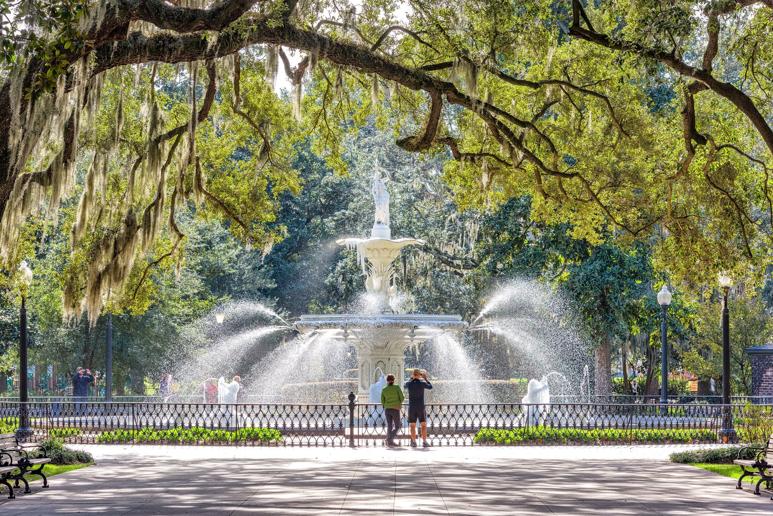 Fountain with ornate design in a park, surrounded by trees and Spanish moss, with people admiring and photographing it.