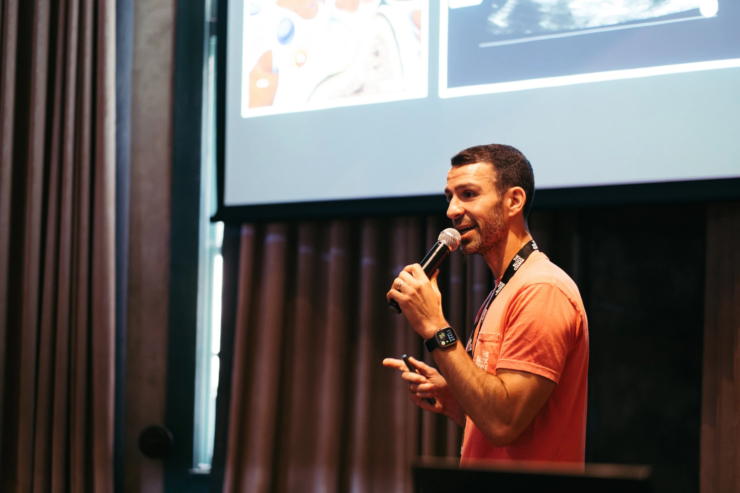 Man wearing an orange shirt and smartwatch speaking into a microphone during a presentation, with a large screen displaying informational slides in the background.