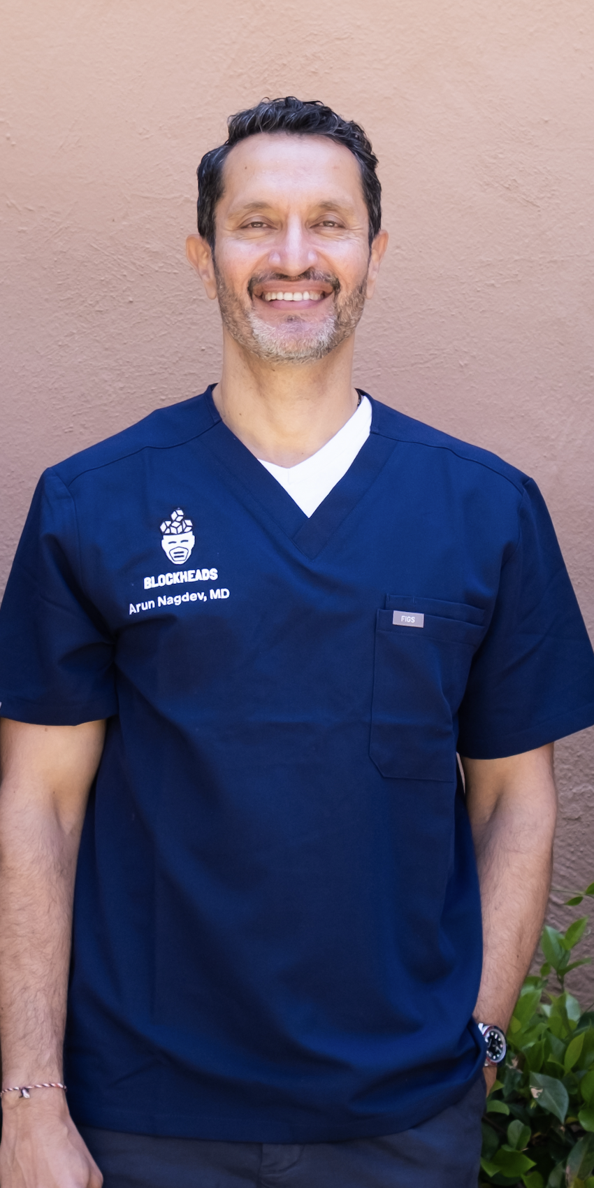 A smiling man wearing a navy scrubs shirt with a logo and text, standing outside against a pink wall.